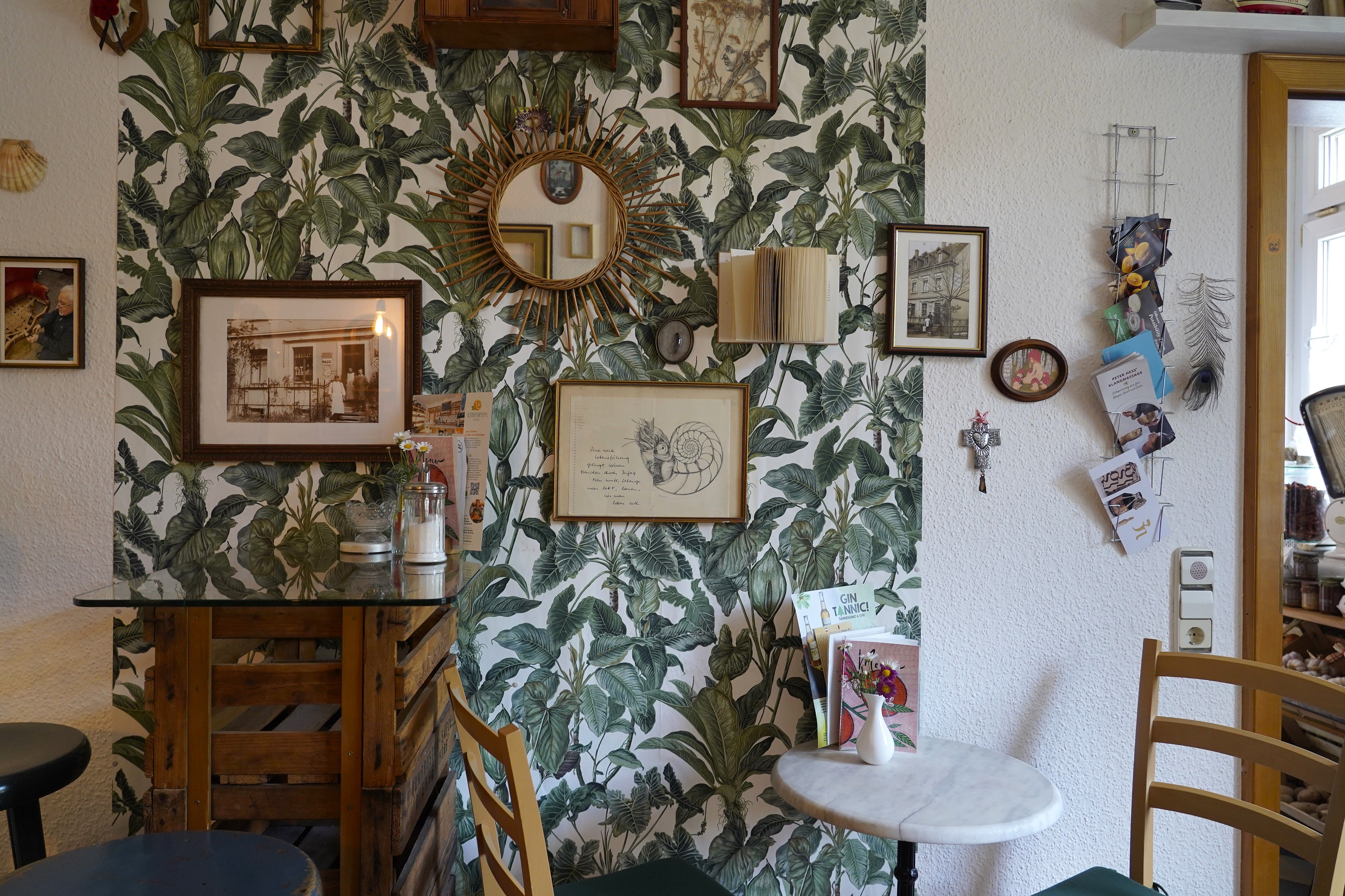 Gemütliche Café-Ecke mit Pflanzen-Tapete, Spiegel, Bildern und Dekorationen an der Wand. Ein kleiner Tisch mit Vase und Stühlen ergänzt das Ambiente.