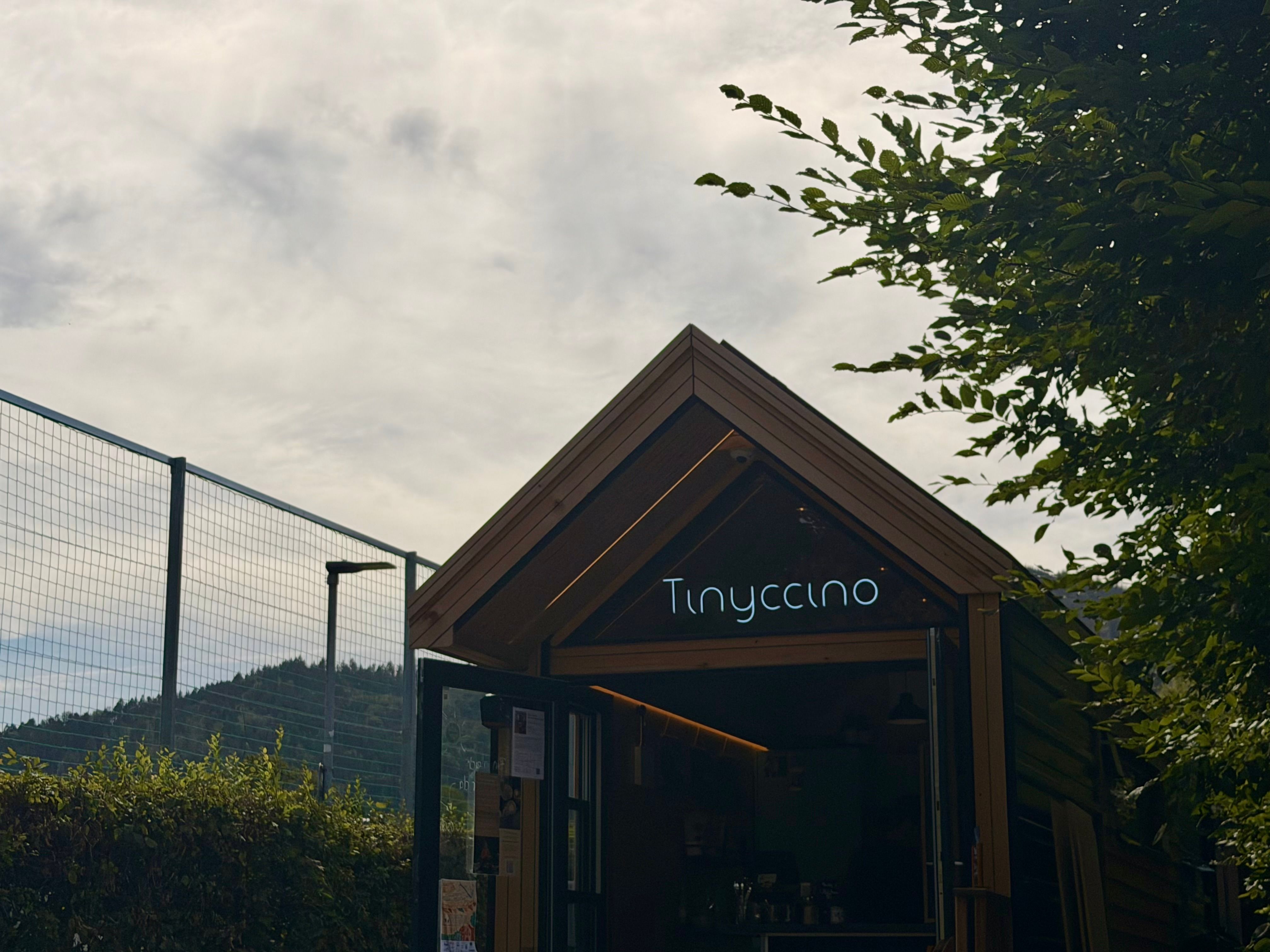 Das kleine Café Tinyccino steht unter einem bewölkten Himmel, eingerahmt von Bäumen und einem Zaun.
