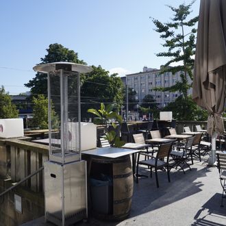 Sonnige Restaurantterrasse mit leeren Tischen und Stühlen, Heizstrahler und Sonnenschirm, umgeben von städtischen Gebäuden und Bäumen.