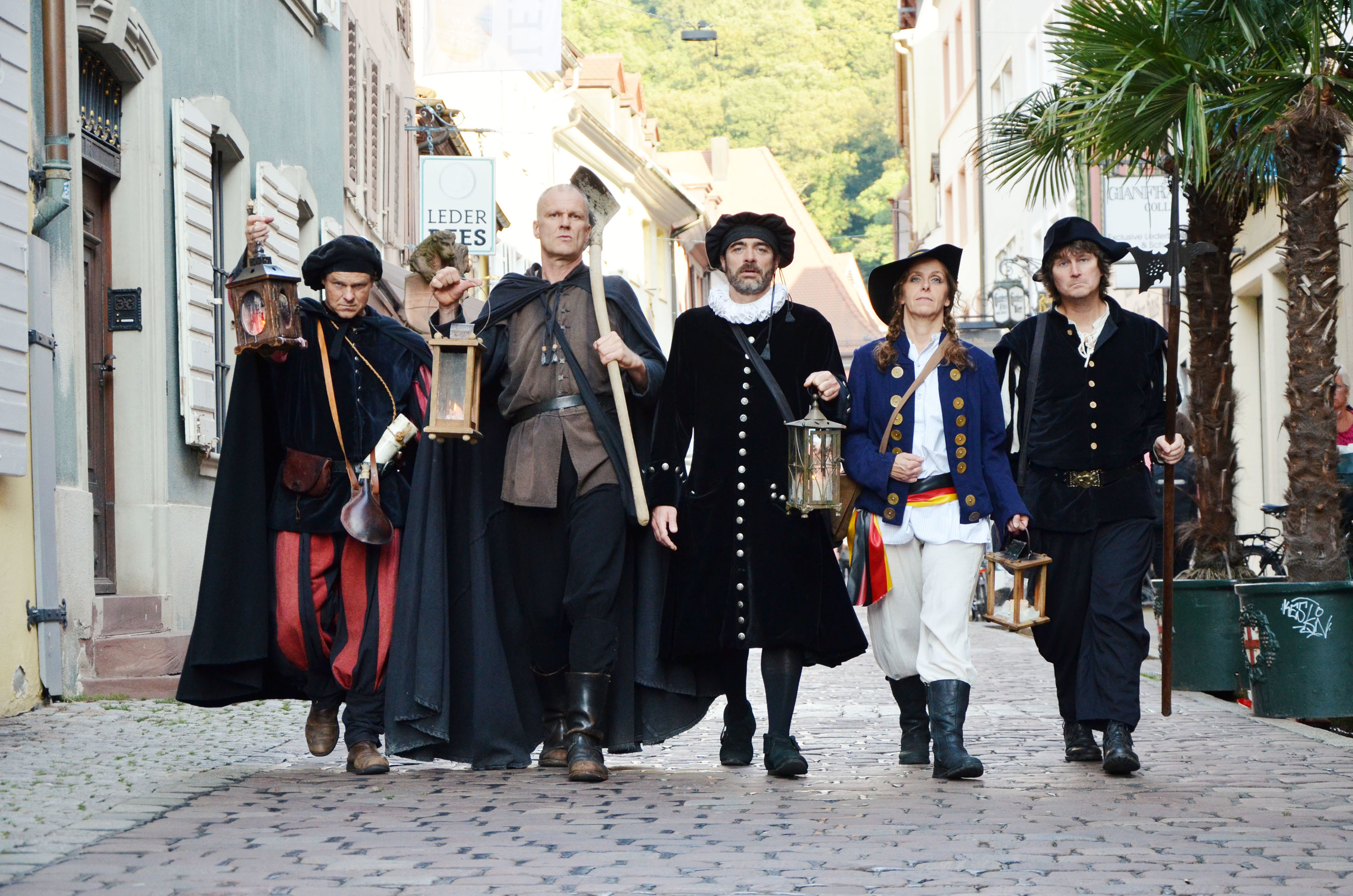 Fünf Personen in historischen Kostümen mit Laternen und Werkzeugen gehen durch eine gepflasterte Altstadtstraße.