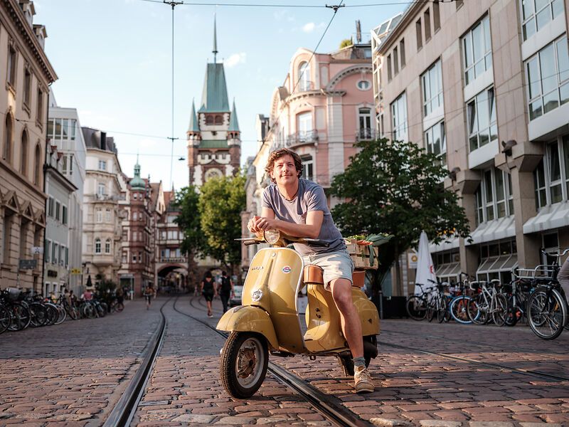 Mann auf gelber Vespa in einer Altstadtstraße mit historischen Gebäuden und Straßenbahnschienen.