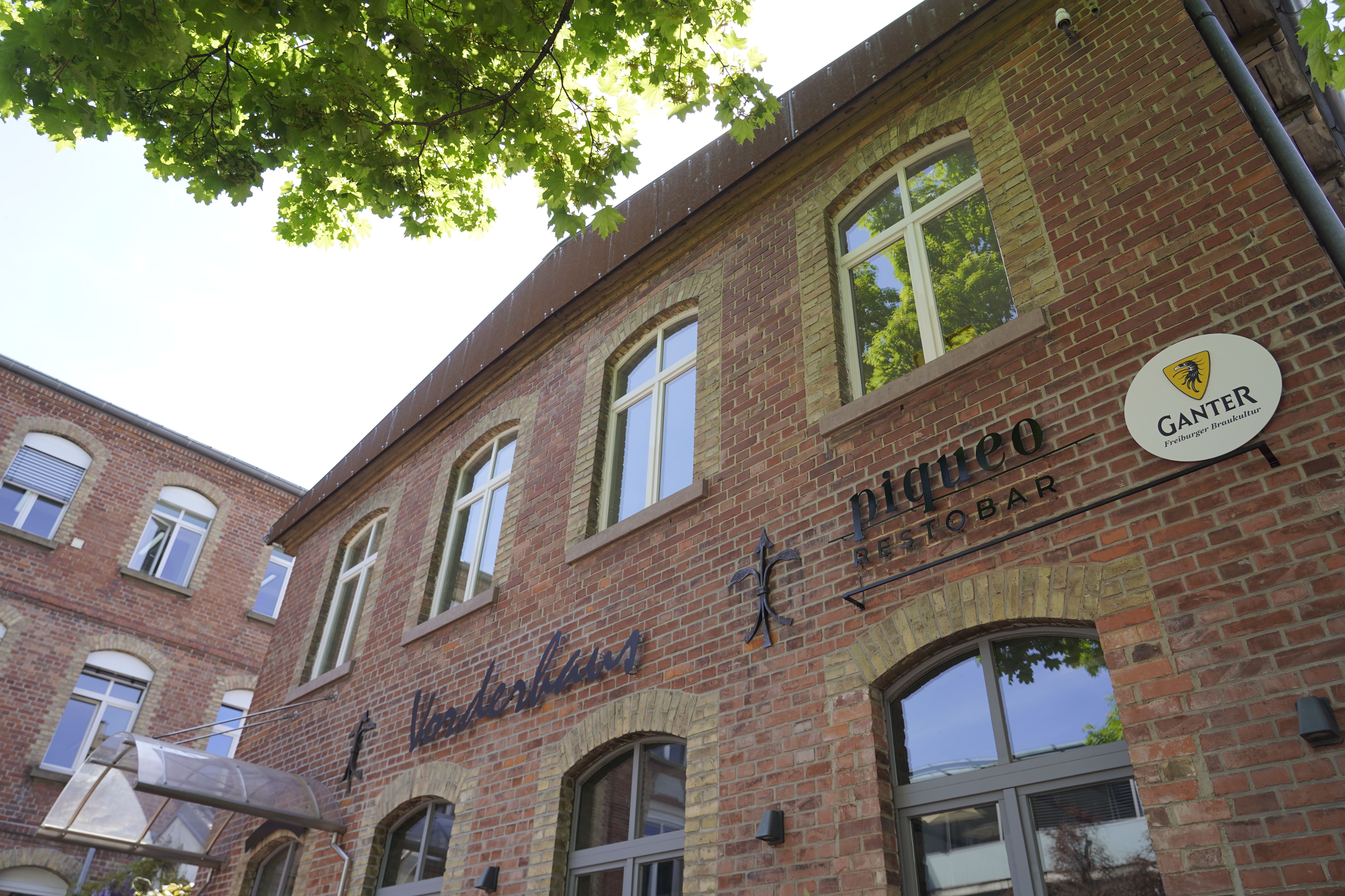 Backsteinfassade des piqueo Restobars im Vorderhaus, umgeben von grünen Blättern. Ein Ganter-Schild ist sichtbar.
