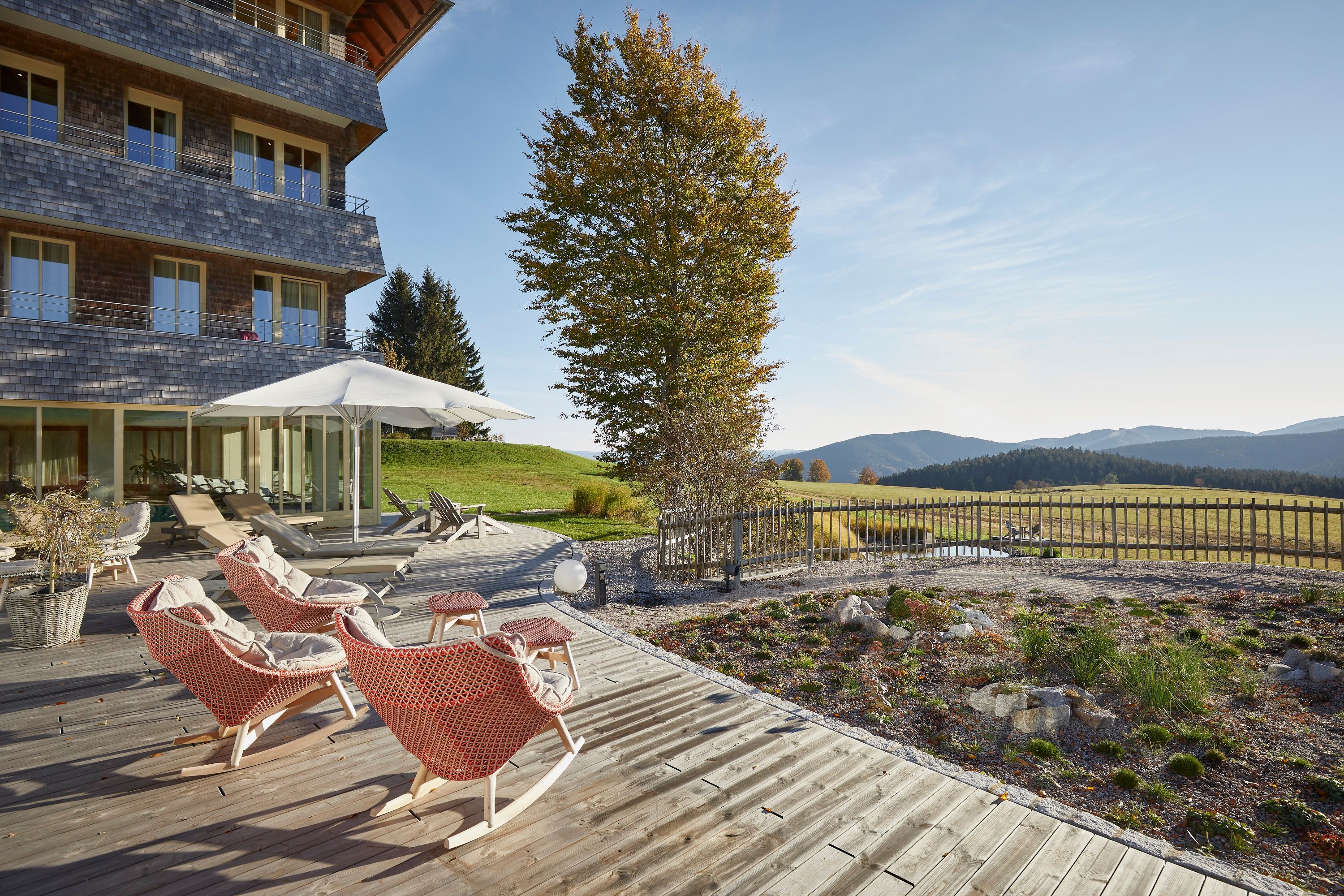 Holzterrasse mit Liegestühlen und Sonnenschirm, umgeben von grüner Landschaft und Bergen im Hintergrund.