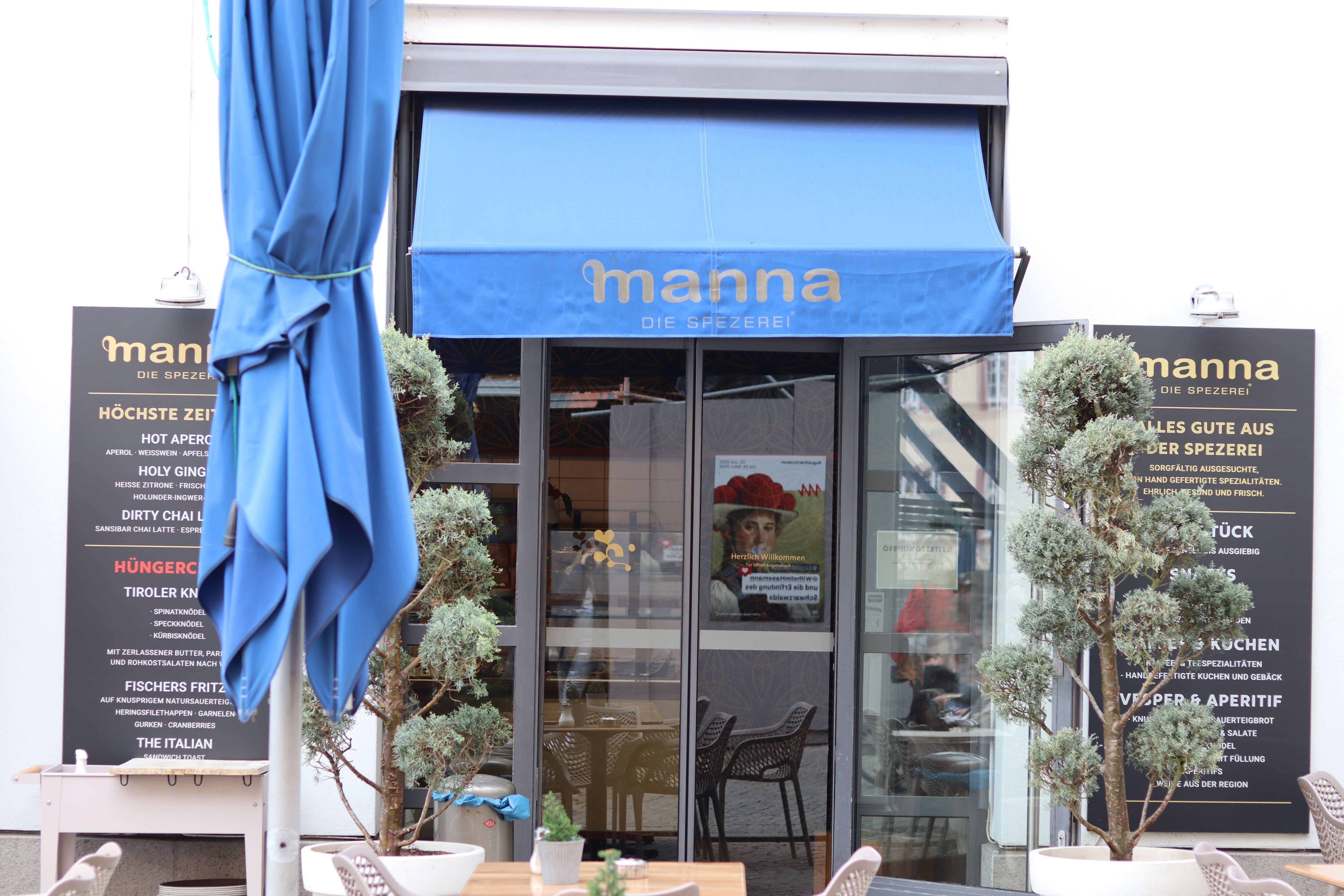 Eingang eines Cafés namens "manna" mit blauer Markise. Zwei Pflanzen flankieren den Eingang, daneben sind Tafeln mit Speisenangeboten.