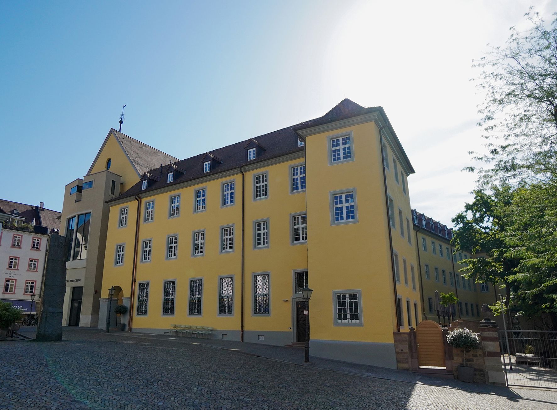 Das Augustinermuseum in Freiburg zeigt sich mit seiner gelben Fassade und blauen Fenstern im Sonnenlicht. Der gepflasterte Vorplatz ist leer.