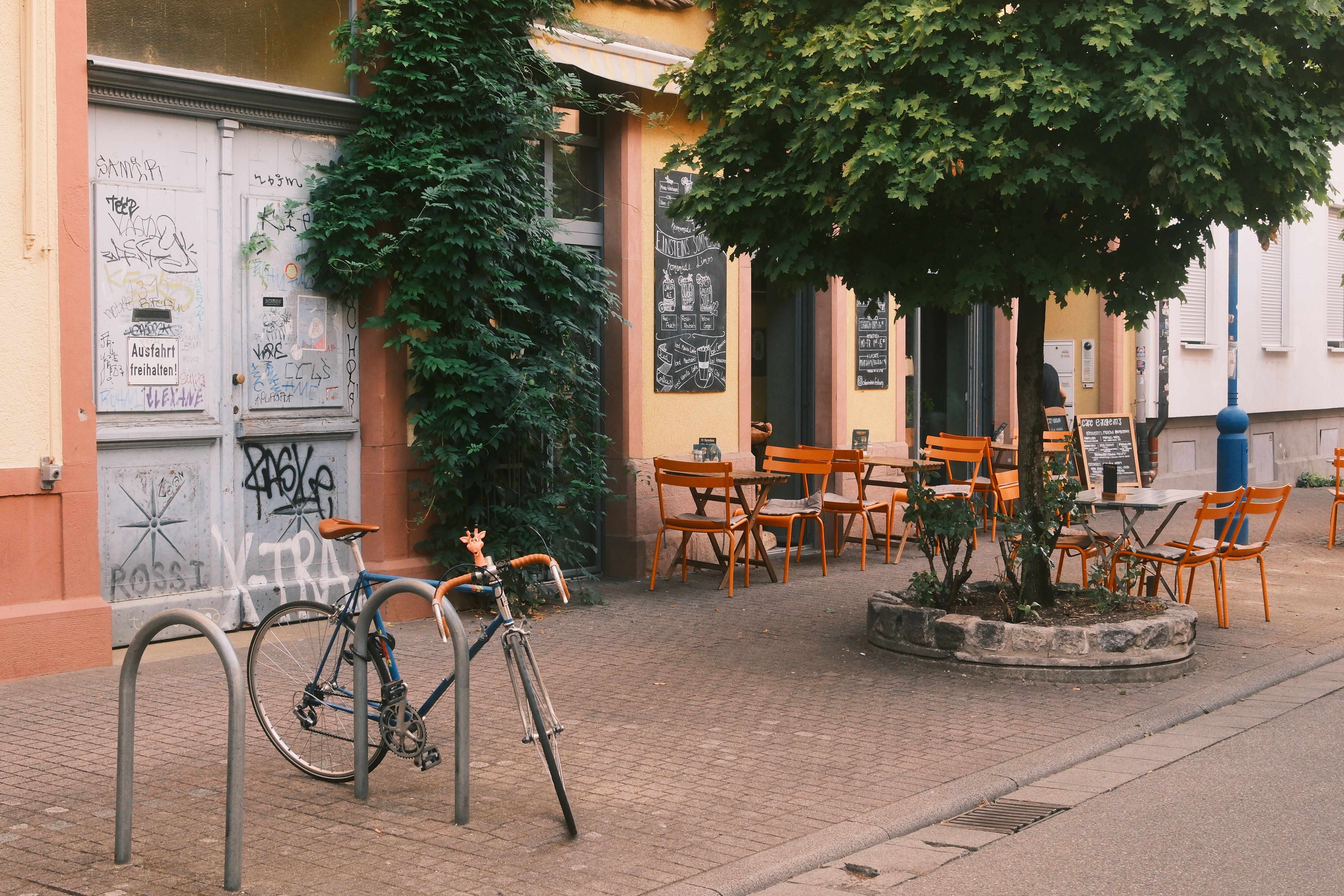Ein gemütliches Straßencafé mit orangen Stühlen und Tischen unter einem Baum. Ein Fahrrad ist an einem Ständer angeschlossen.