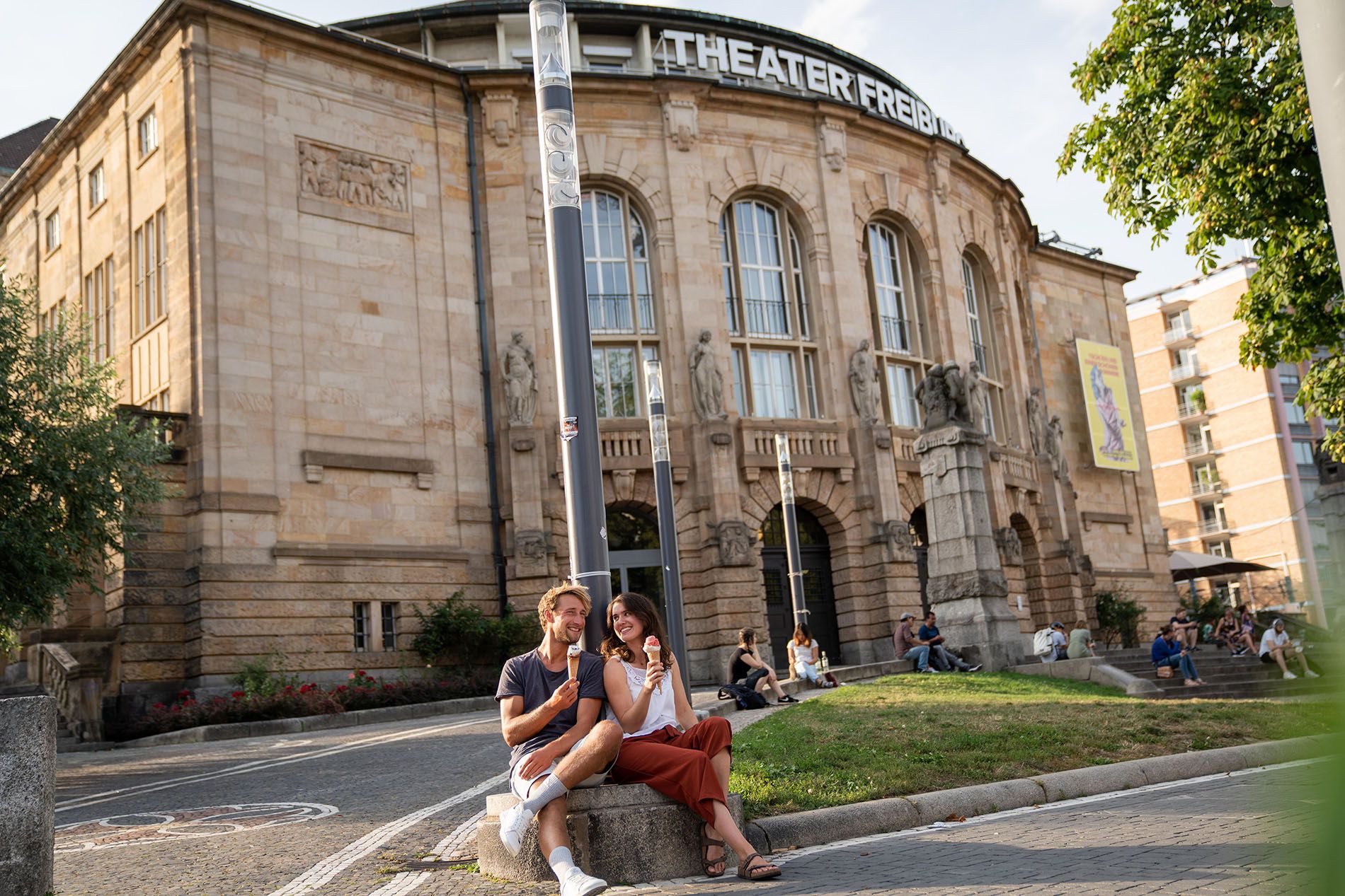 Blick auf das Theater Freiburg, im Vordergrund sitzt ein paar und isst Eis