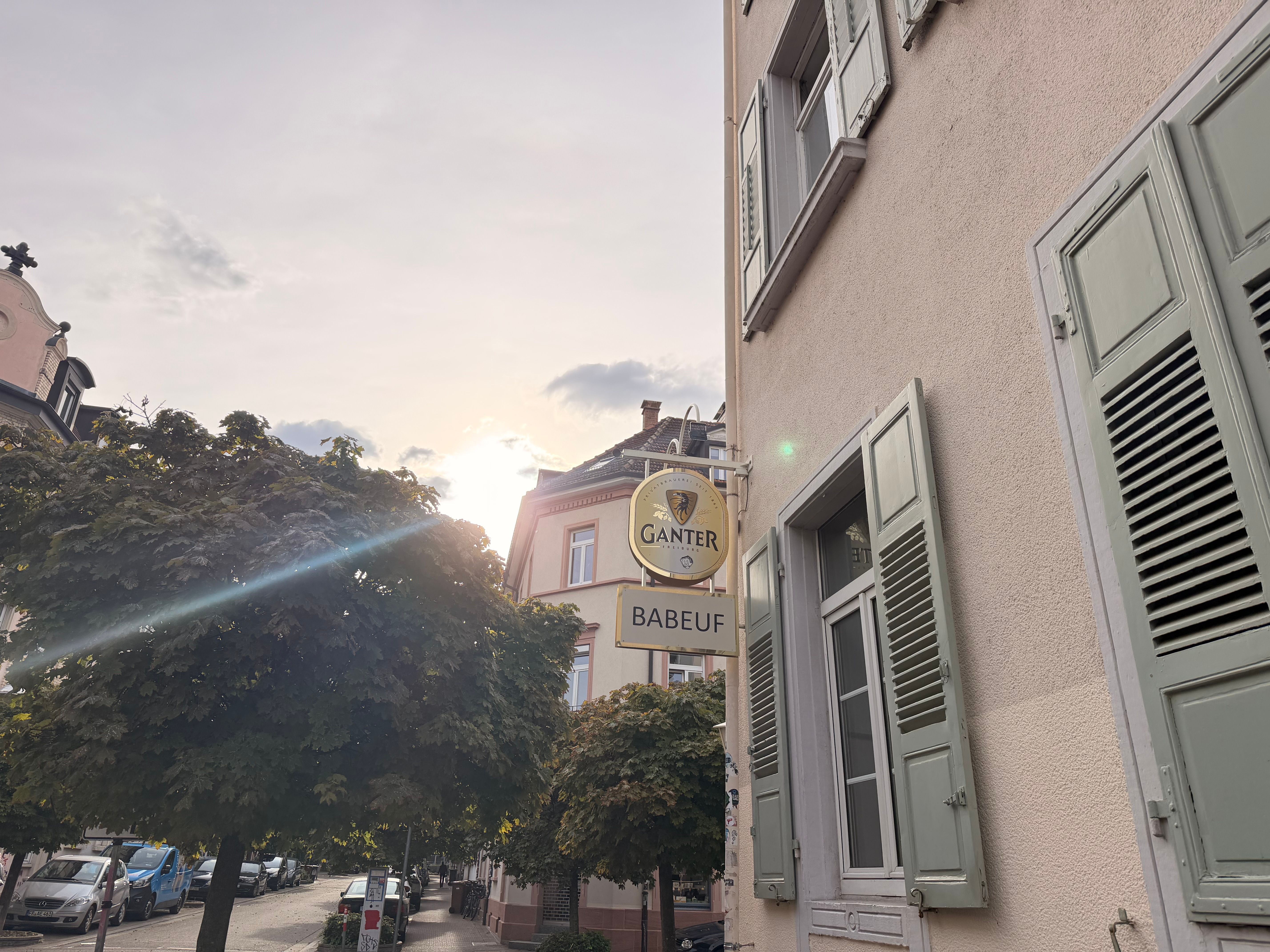Straßenszene mit dem Schild „Babeuf“ und dem „Ganter“-Wirtshausschild an einem Haus mit grünen Fensterläden im Abendlicht.