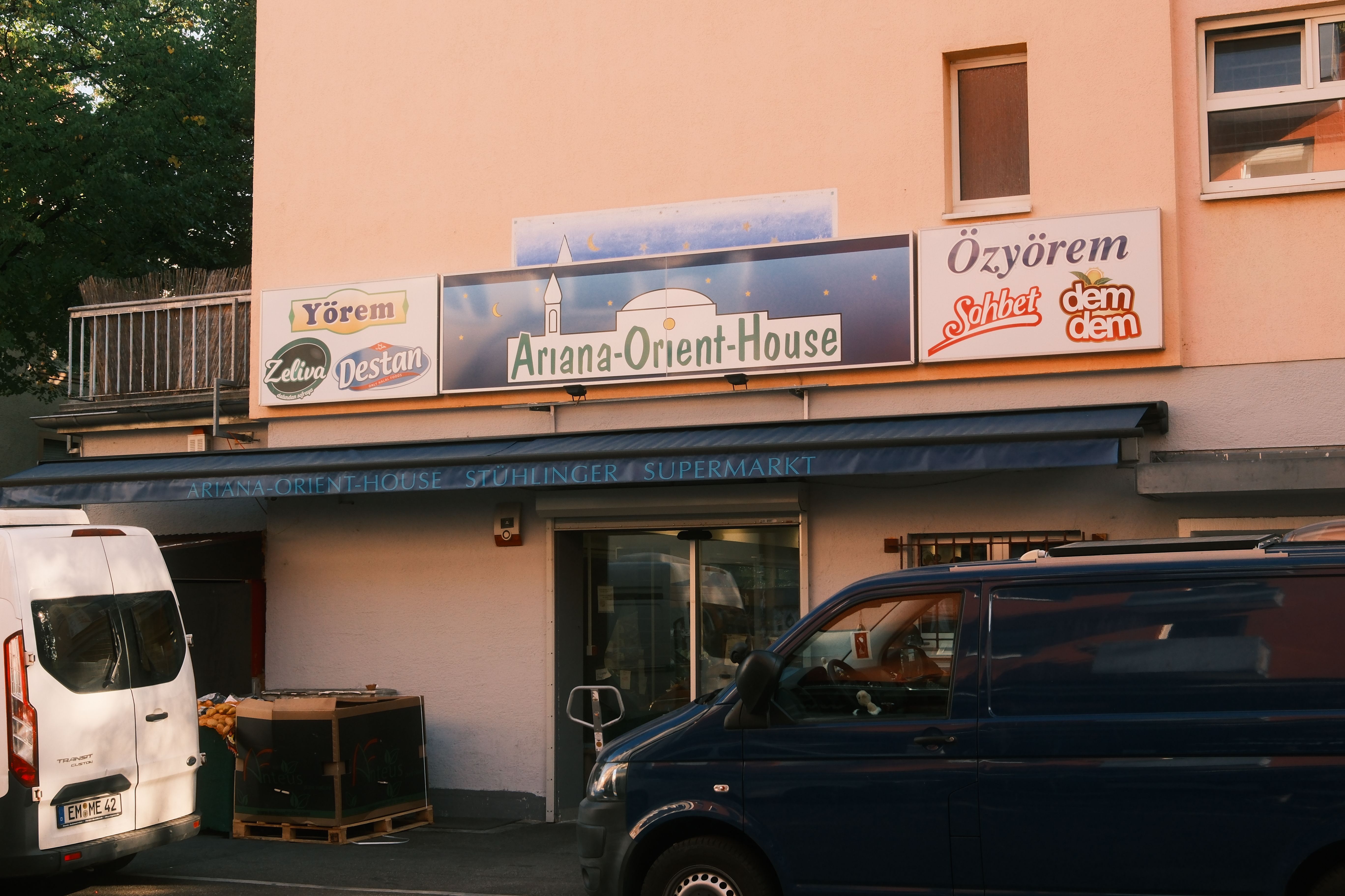 Der Eingang des Ariana-Orient-House Supermarkts im Stadtteil Stühlinger mit verschiedenen Werbeschildern und parkenden Autos davor.