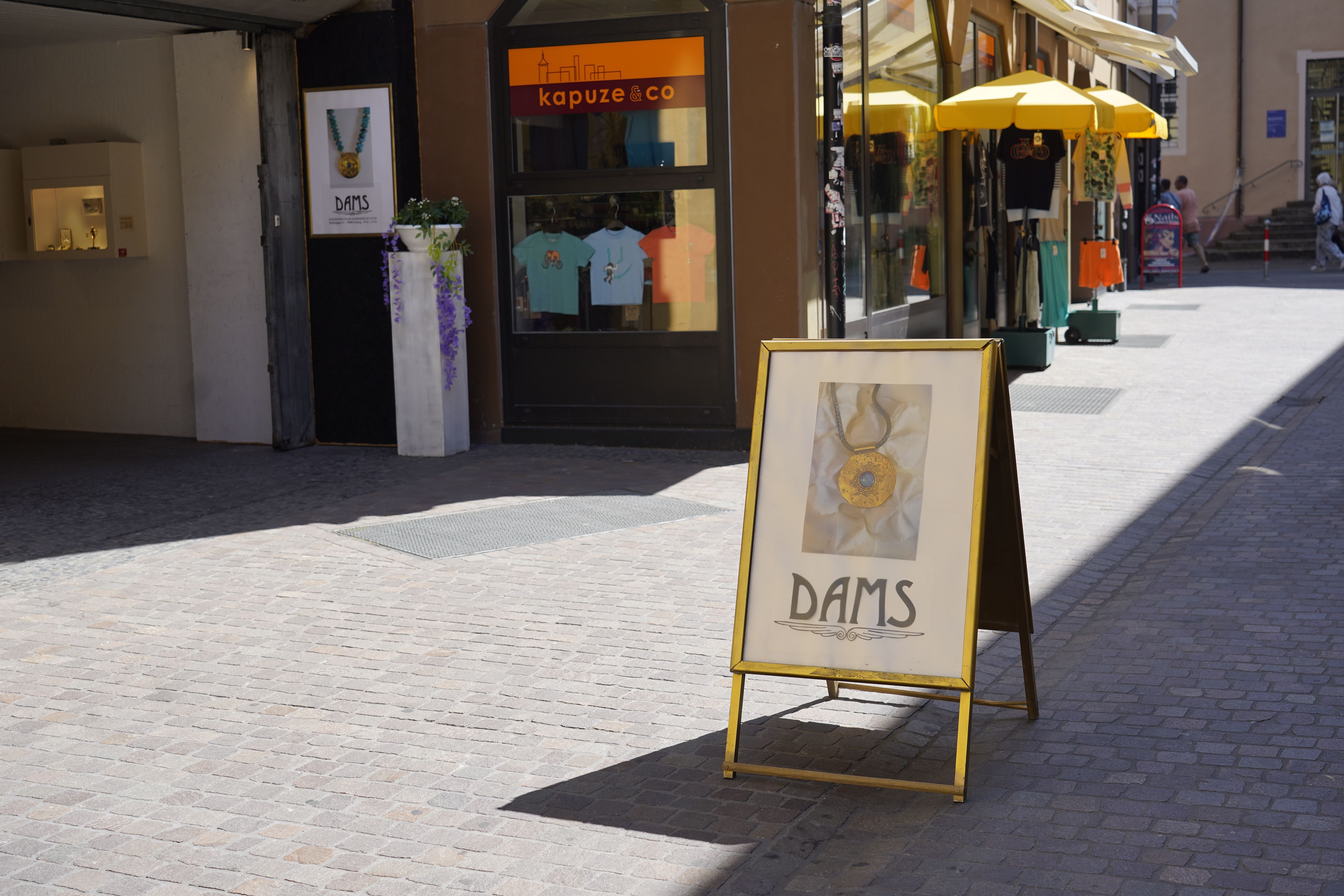Ein Werbeschild für das Schmuckgeschäft Dams steht auf einer gepflasterten Straße. Im Hintergrund sind Geschäfte und Passanten zu sehen.
