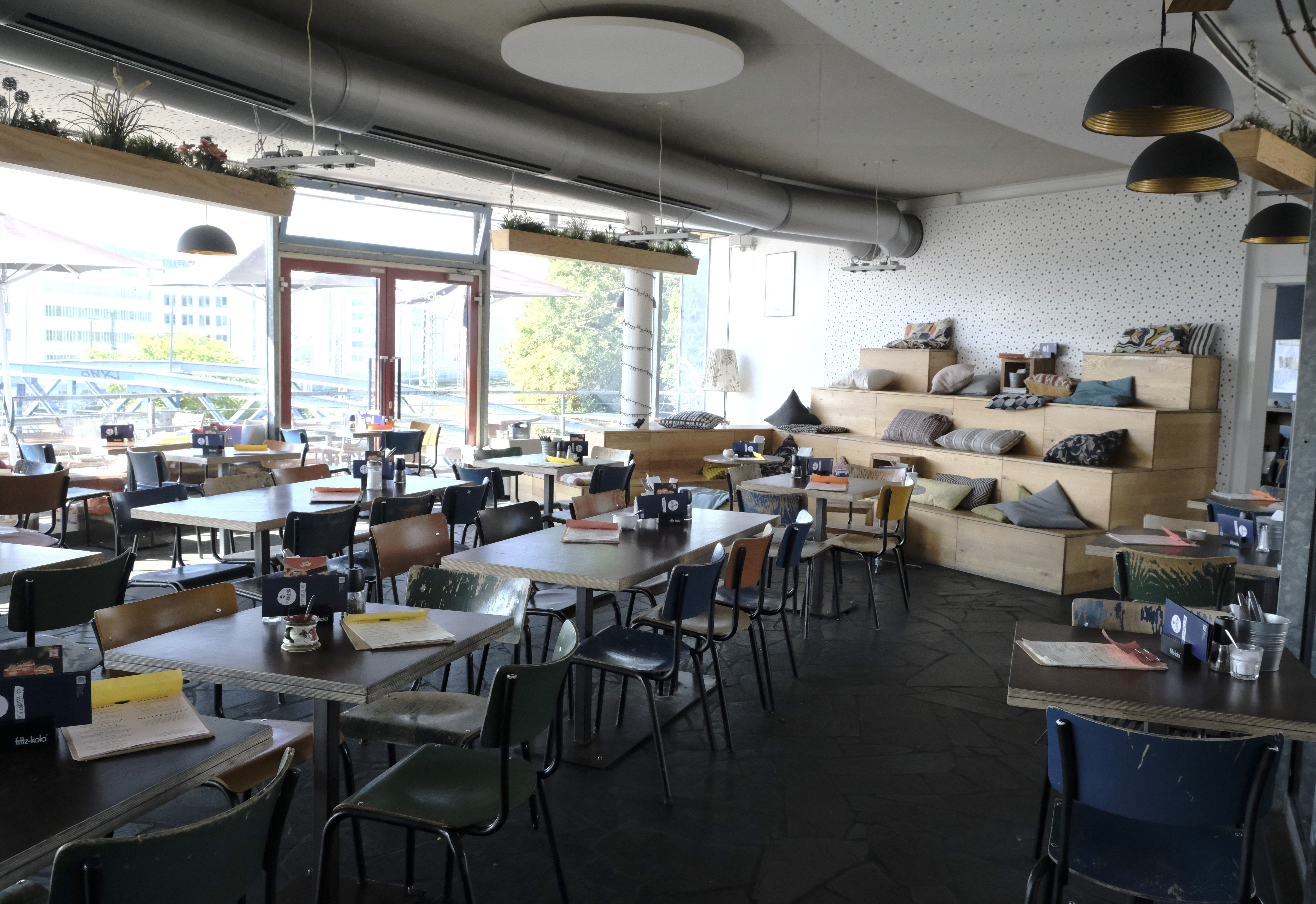Modernes Café mit Holztischen, bunten Stühlen und einer Sitztreppe mit Kissen. Große Fenster bieten Blick nach draußen.