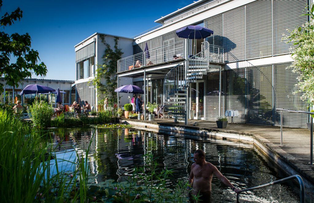 Wellnessbereich mit Teich, Sonnenschirmen und Menschen. Moderne Architektur mit Terrasse und Wendeltreppe. Entspannte Atmosphäre im Freien.