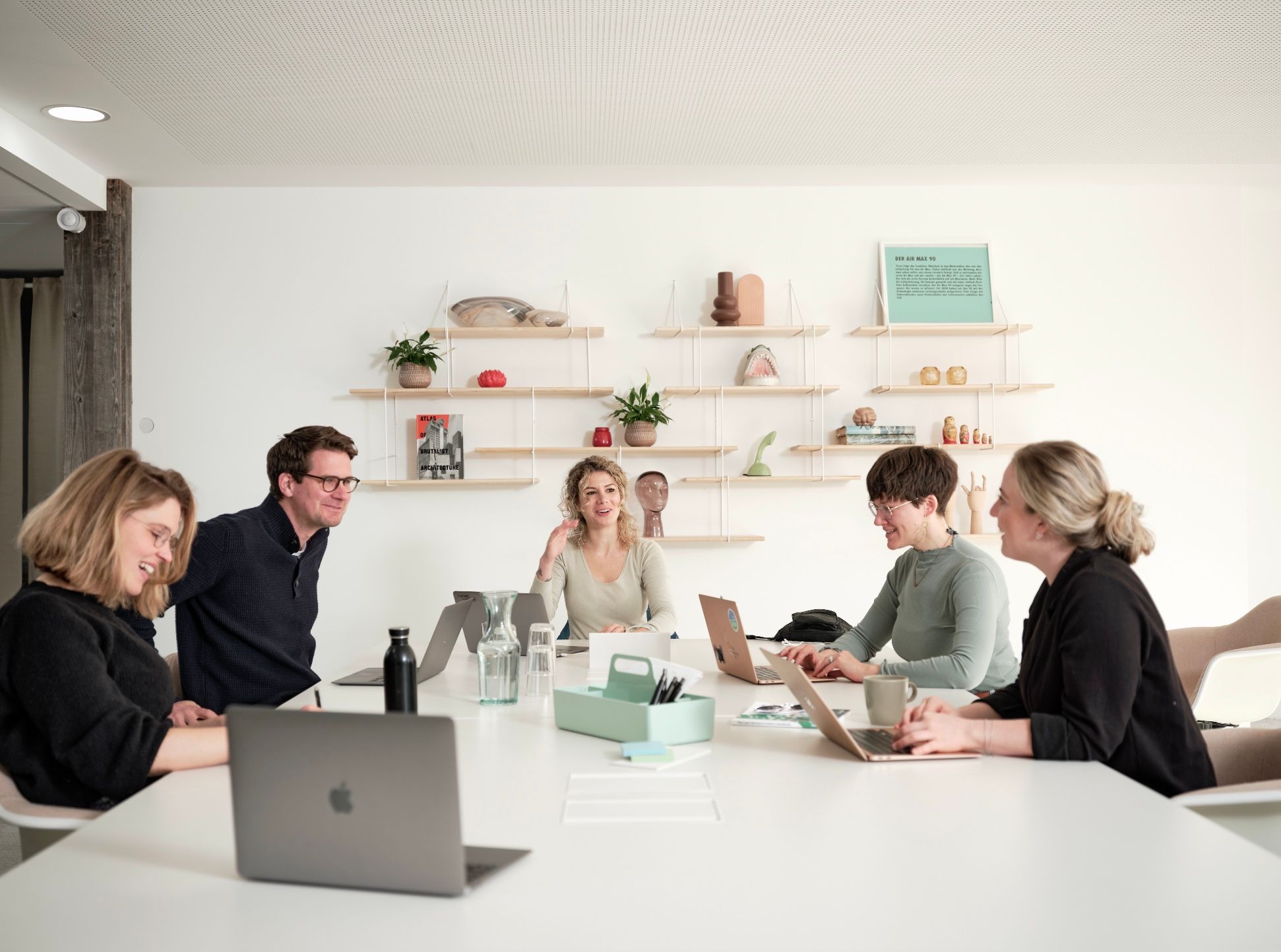 Ein Team von fünf Personen sitzt in einem modernen Büro an einem Tisch und arbeitet gemeinsam an Laptops. Regale mit Pflanzen im Hintergrund.