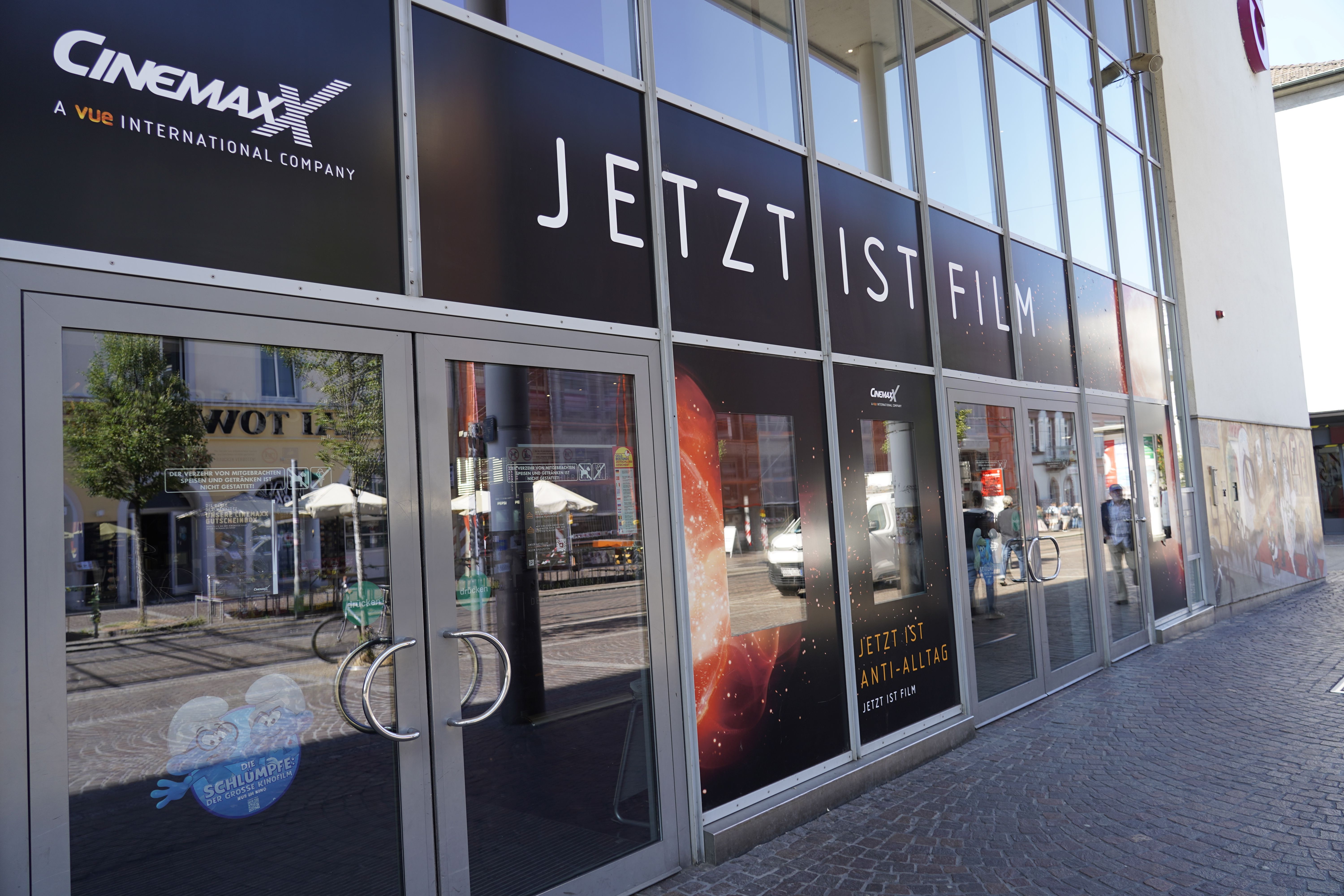 Eingang des Cinemaxx Kinos in Freiburg mit Glasfassade und Werbeplakaten. Aufschrift: 'Jetzt ist Film'. Reflektionen von Gebäuden und Passanten sichtbar.