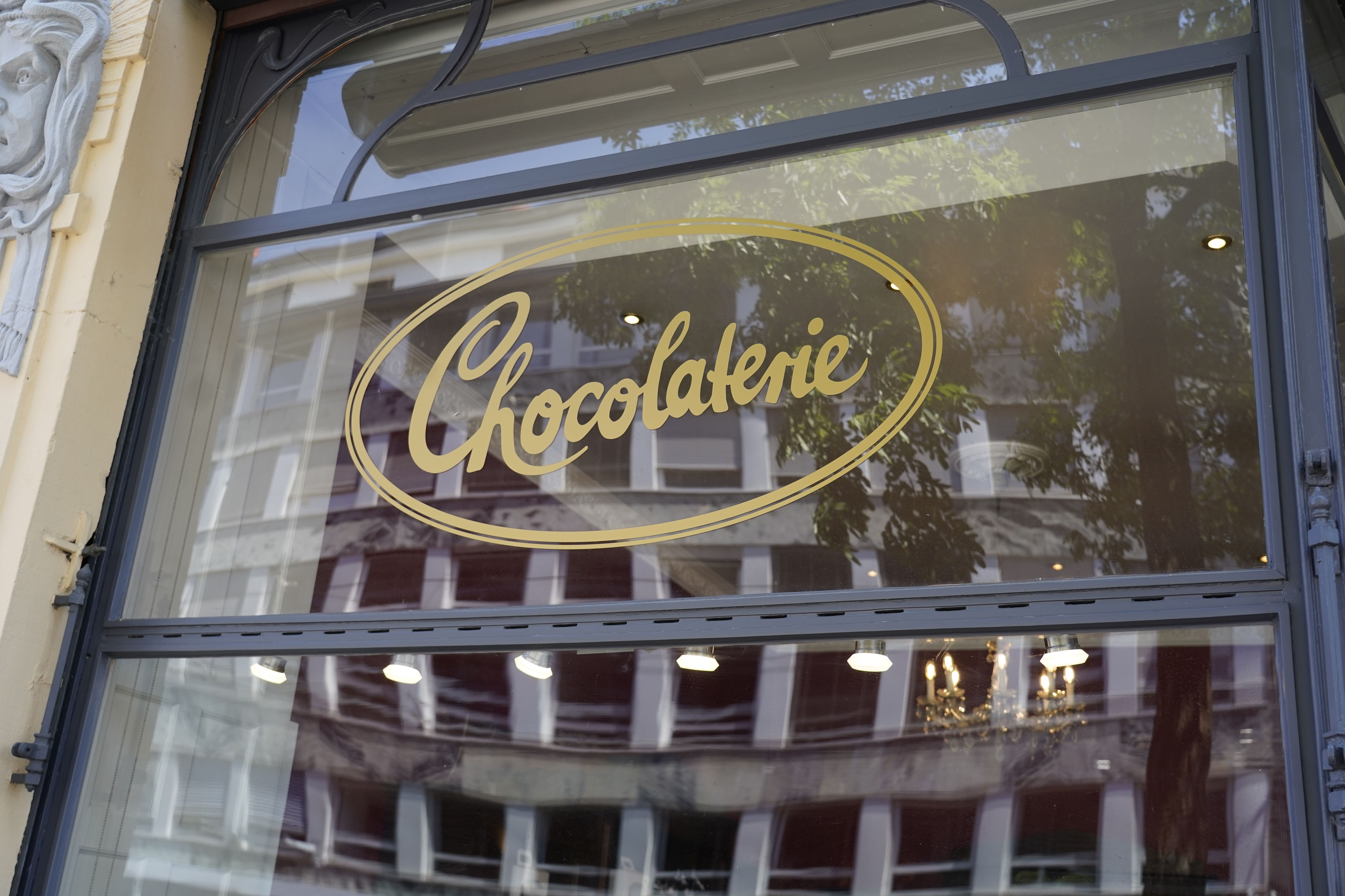 Schaufenster einer Chocolaterie mit goldener Schrift auf Glas. Im Hintergrund sind Bäume und ein Kronleuchter zu sehen.