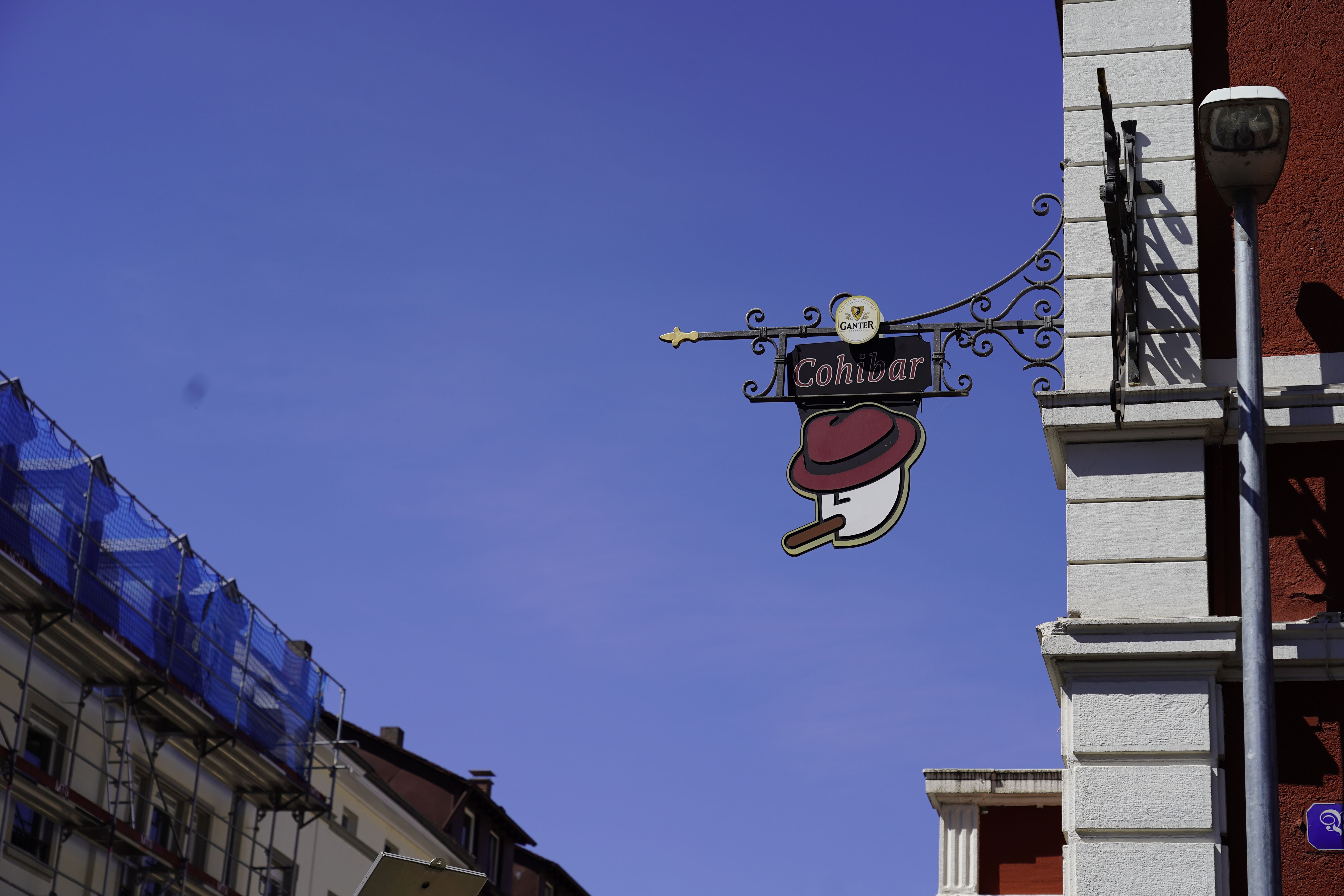 Schild der Cohibar mit stilisiertem Gesicht, Hut und Zigarre an einer Hauswand.