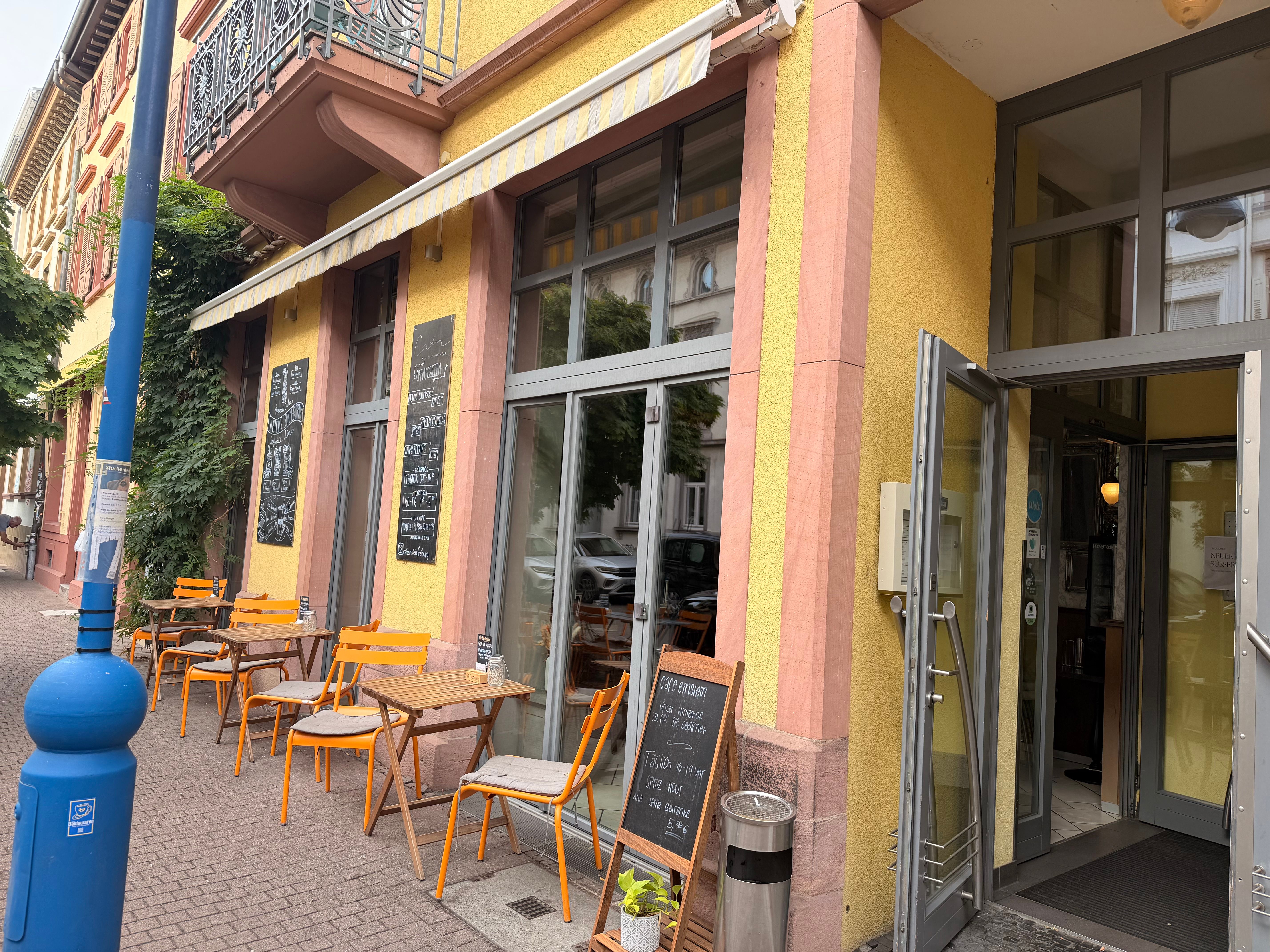 Außenansicht des Café Einstein mit gelber Fassade, offenen Glastüren und Sitzplätzen auf dem Gehweg.