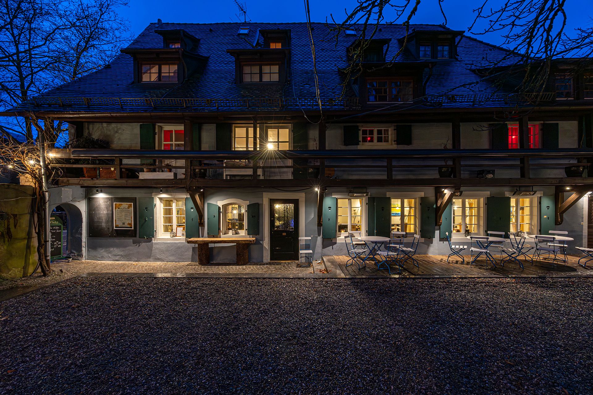 Ein gemütliches Gasthaus mit beleuchteten Fenstern und leeren Tischen im Freien bei Dämmerung. Das Gebäude hat grüne Fensterläden und ein Schindeldach.