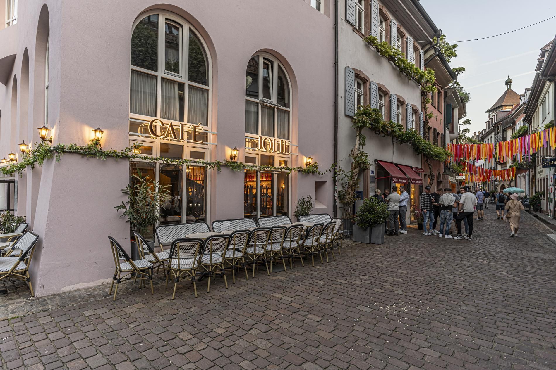 Eine charmante Straße mit einem Café und bunten Bändern, die über die Kopfsteinpflasterstraße gespannt sind. Menschen schlendern und unterhalten sich.
