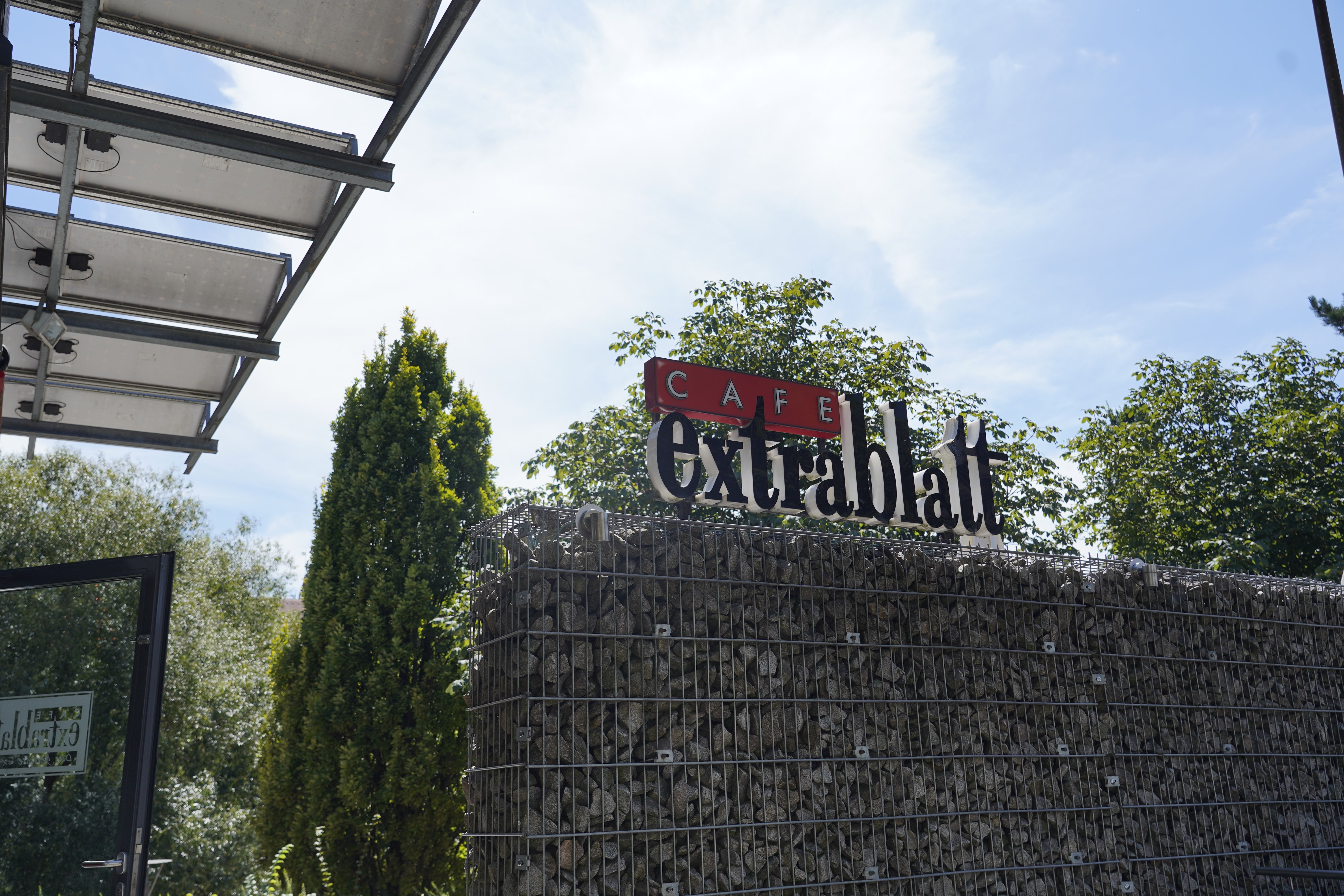 Schild des Café Extrablatt auf einem Steinkorb, umgeben von Bäumen und einem Glasdach.