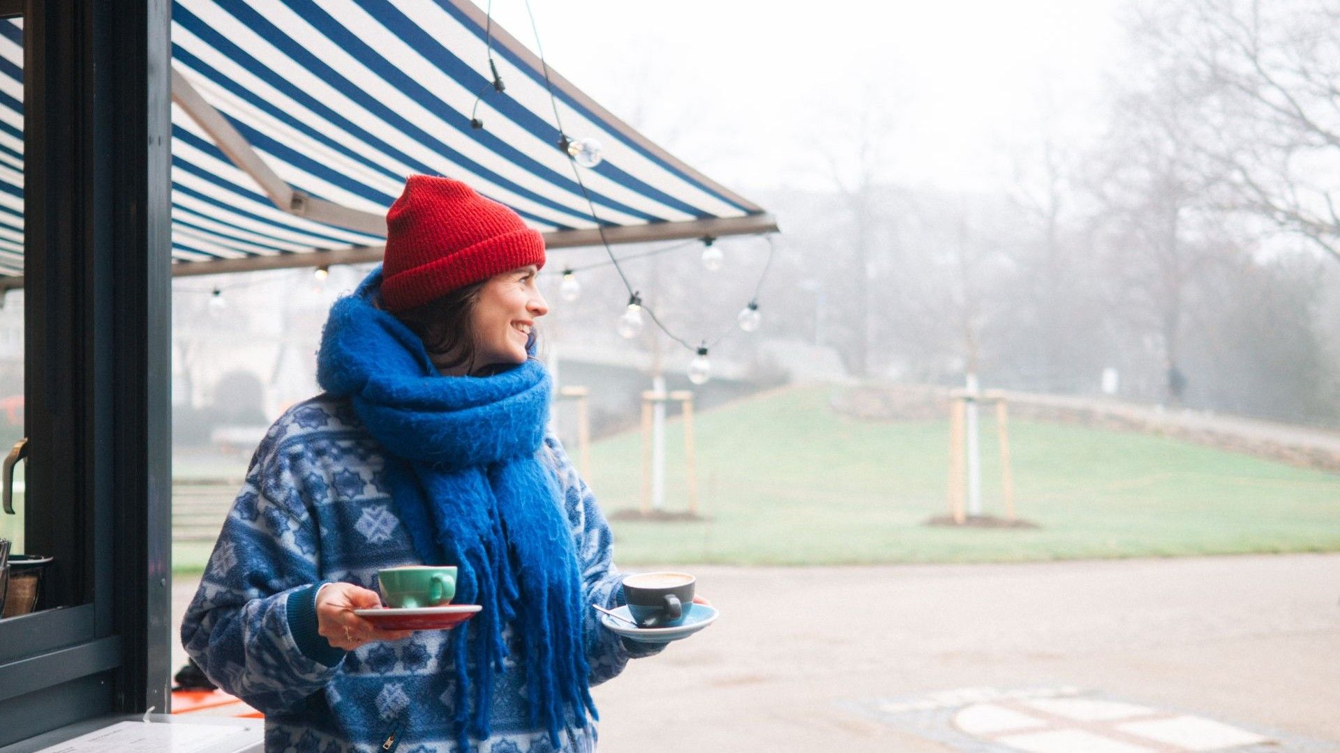 Eine Frau trägt zwei Tassen Kaffee bei diesigem Wetter vor einem Kiosk draußen