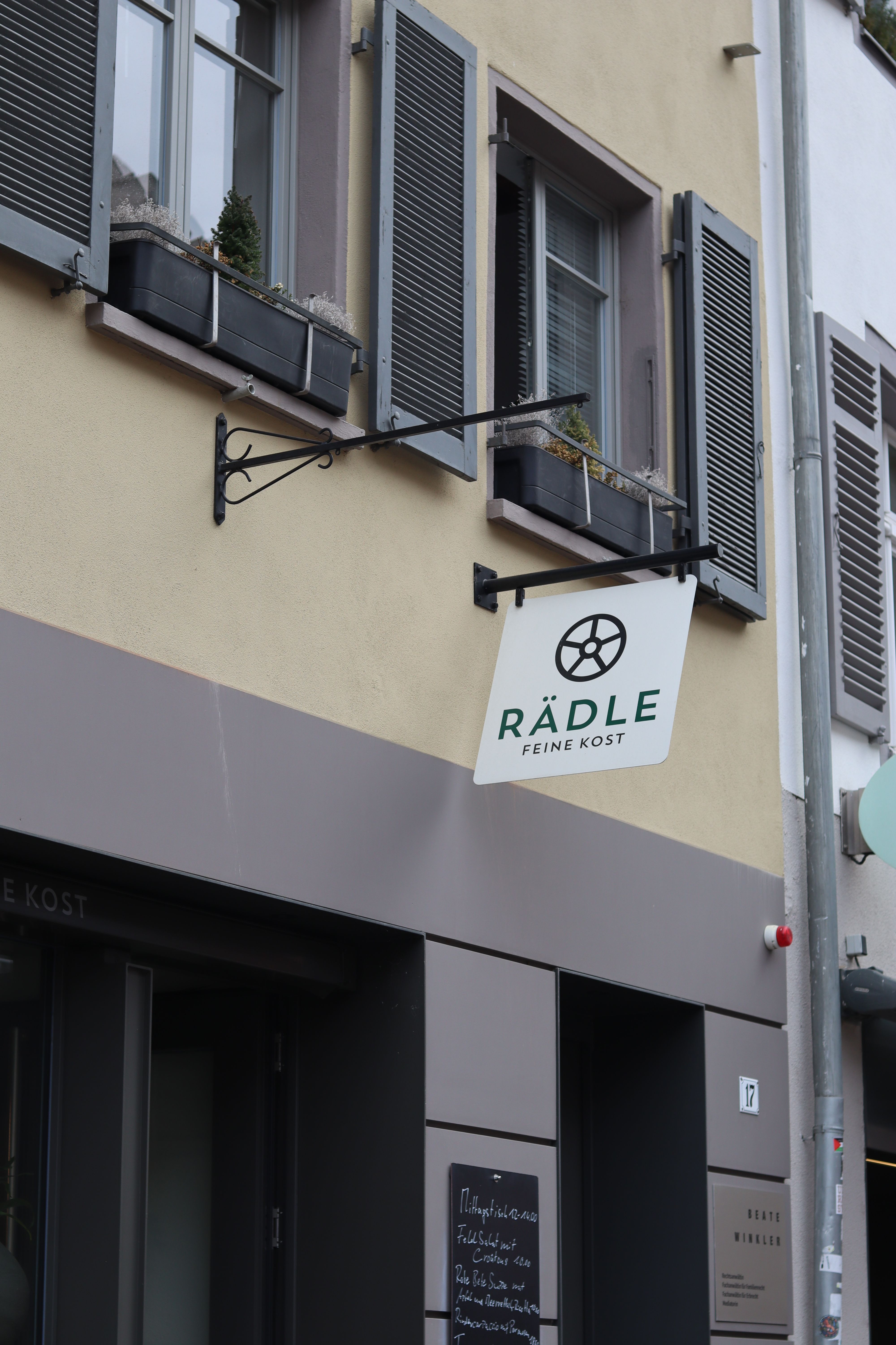 Ein Schild mit der Aufschrift 'RÄDLE FEINE KOST' hängt an einem Gebäude mit grauen Fensterläden und Blumenkästen.