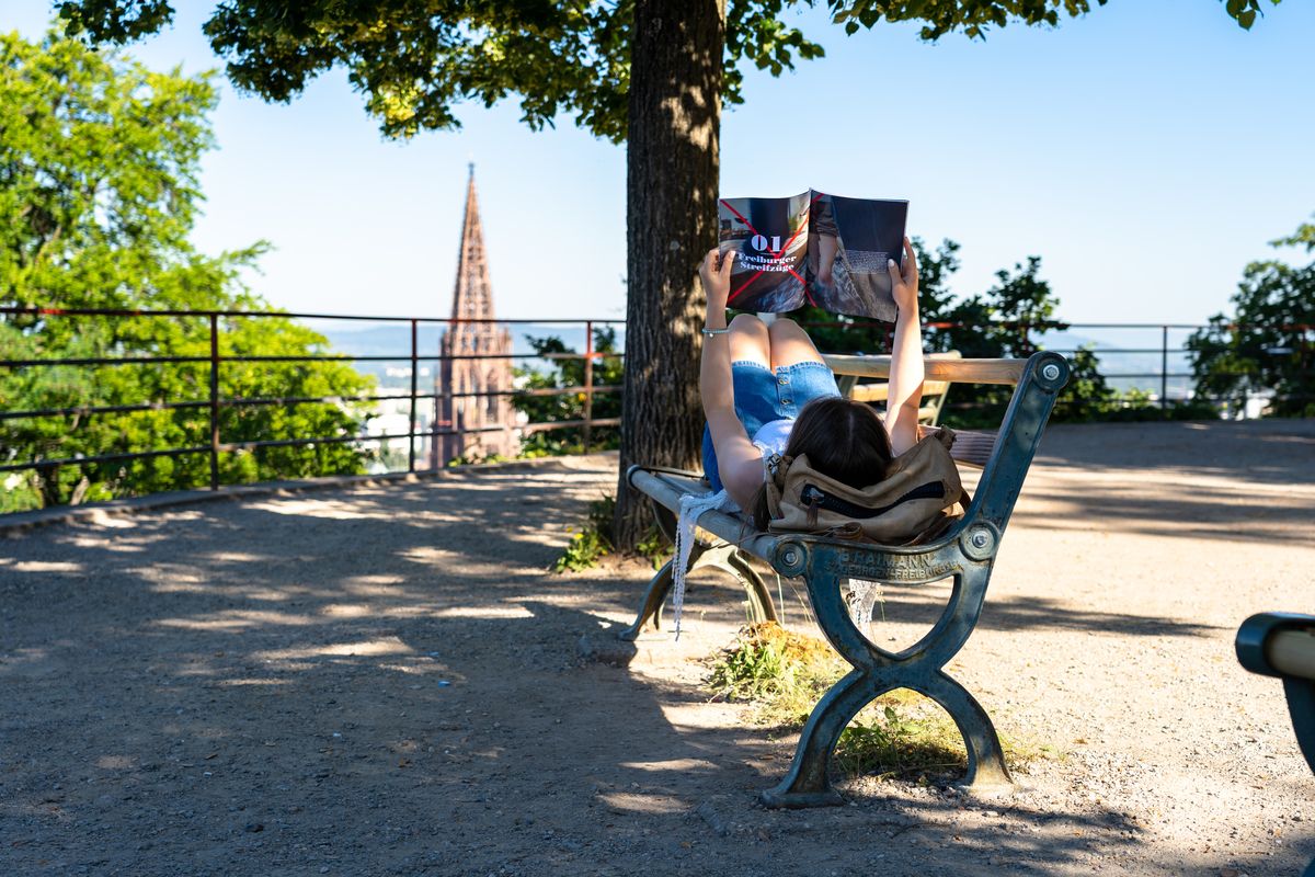 Eine Frau liegt auf einer Bank auf dem Schlossberg und liest eine Zeitschrift. Im Hintergrund sieht man die Aussicht auf Freiburg