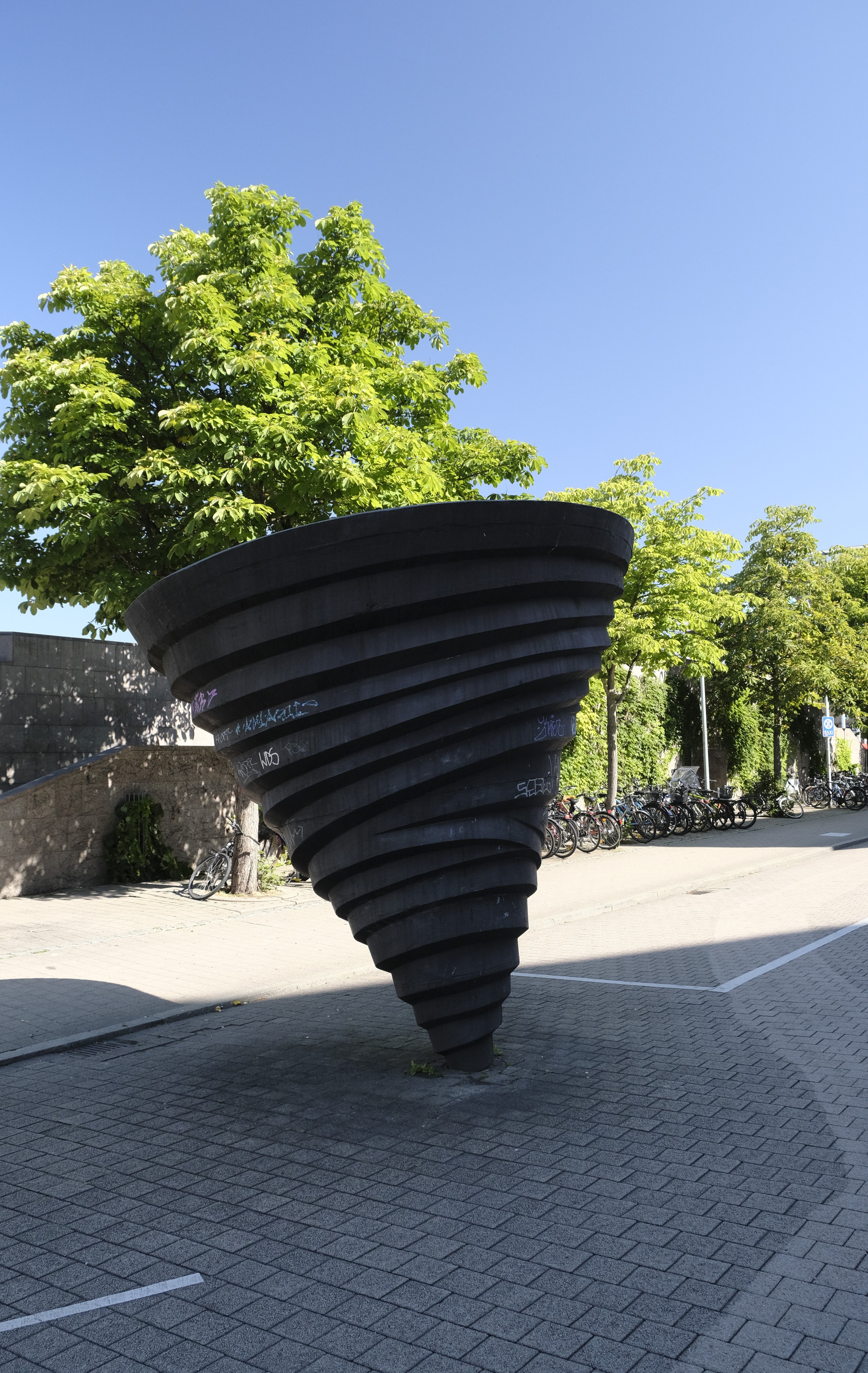 Schwarze, spiralförmige Skulptur auf einem gepflasterten Platz, umgeben von Bäumen und Fahrrädern bei klarem Himmel.