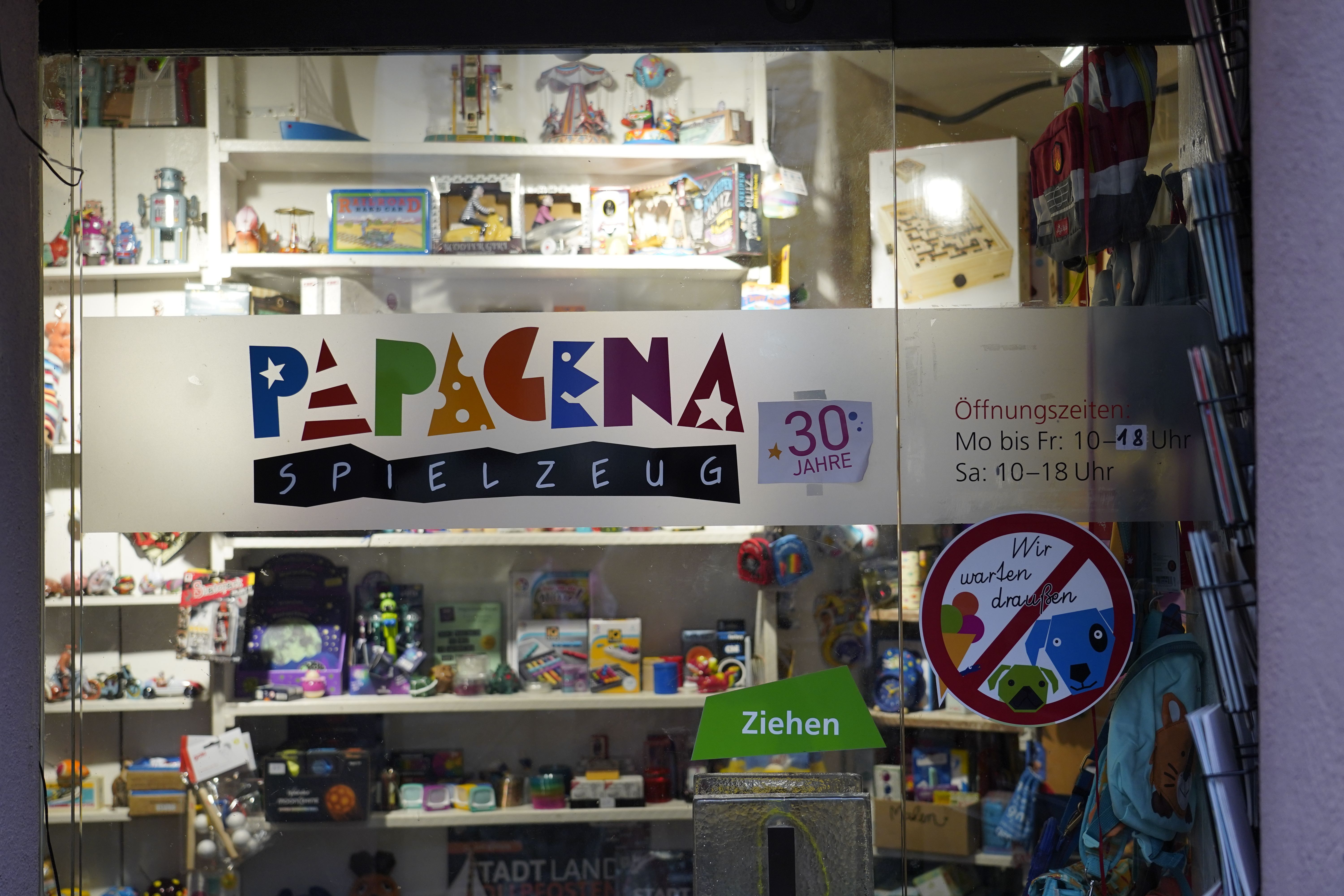 Schaufenster eines Spielzeugladens mit dem bunten Logo 'Papagena Spielzeug'. Im Hintergrund sind verschiedene Spielzeuge zu sehen. Öffnungszeiten sind angegeben.