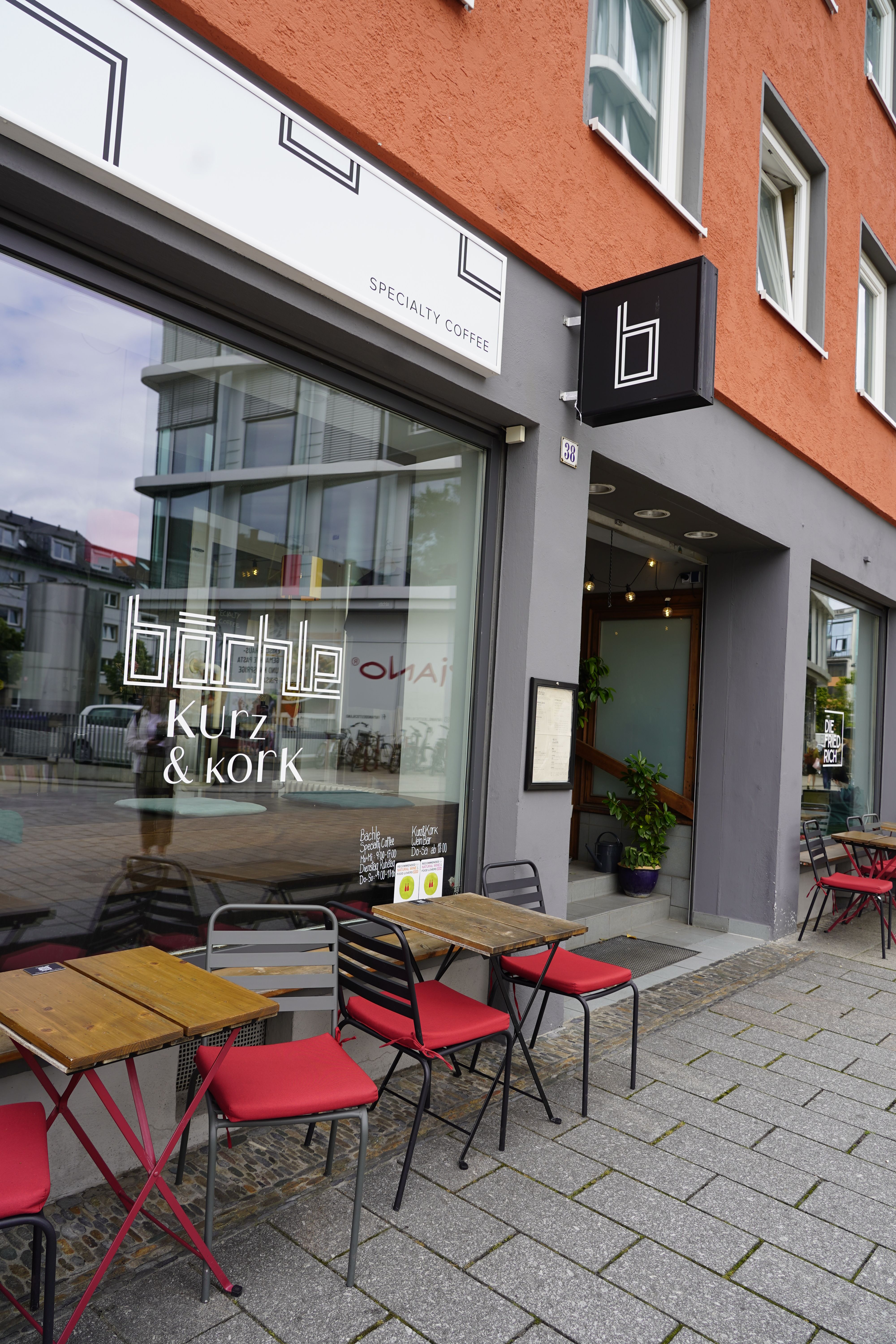Außenansicht des Bächle Specialty Coffee mit roten Stühlen und Holztischen auf dem Gehweg. Das Café hat eine große Fensterfront und ein modernes Design.