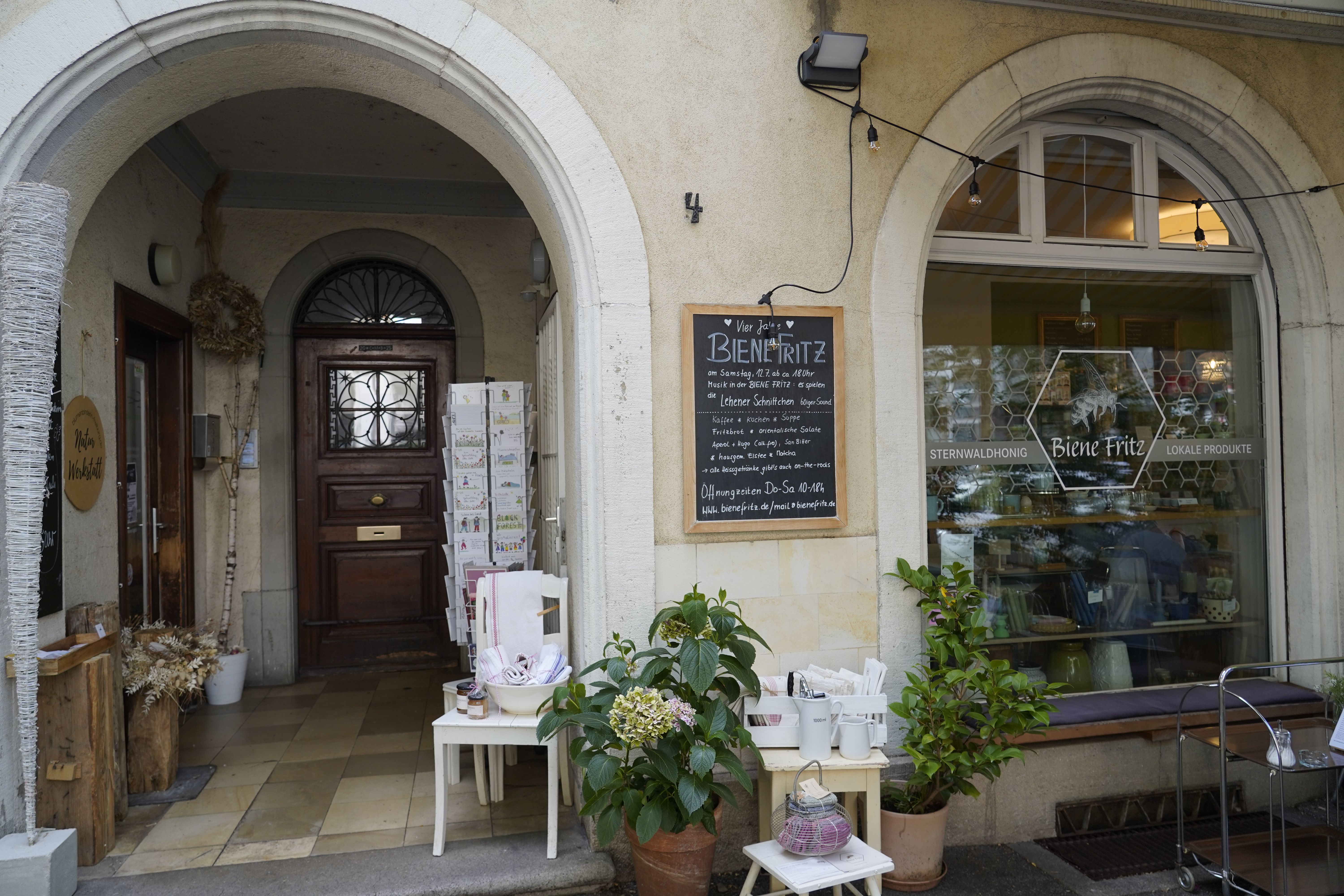 Eingang des Ladens Biene Fritz mit Schaufenster, Pflanzen und einer Tafel mit Informationen. Der Laden bietet lokale Produkte und Honig an.