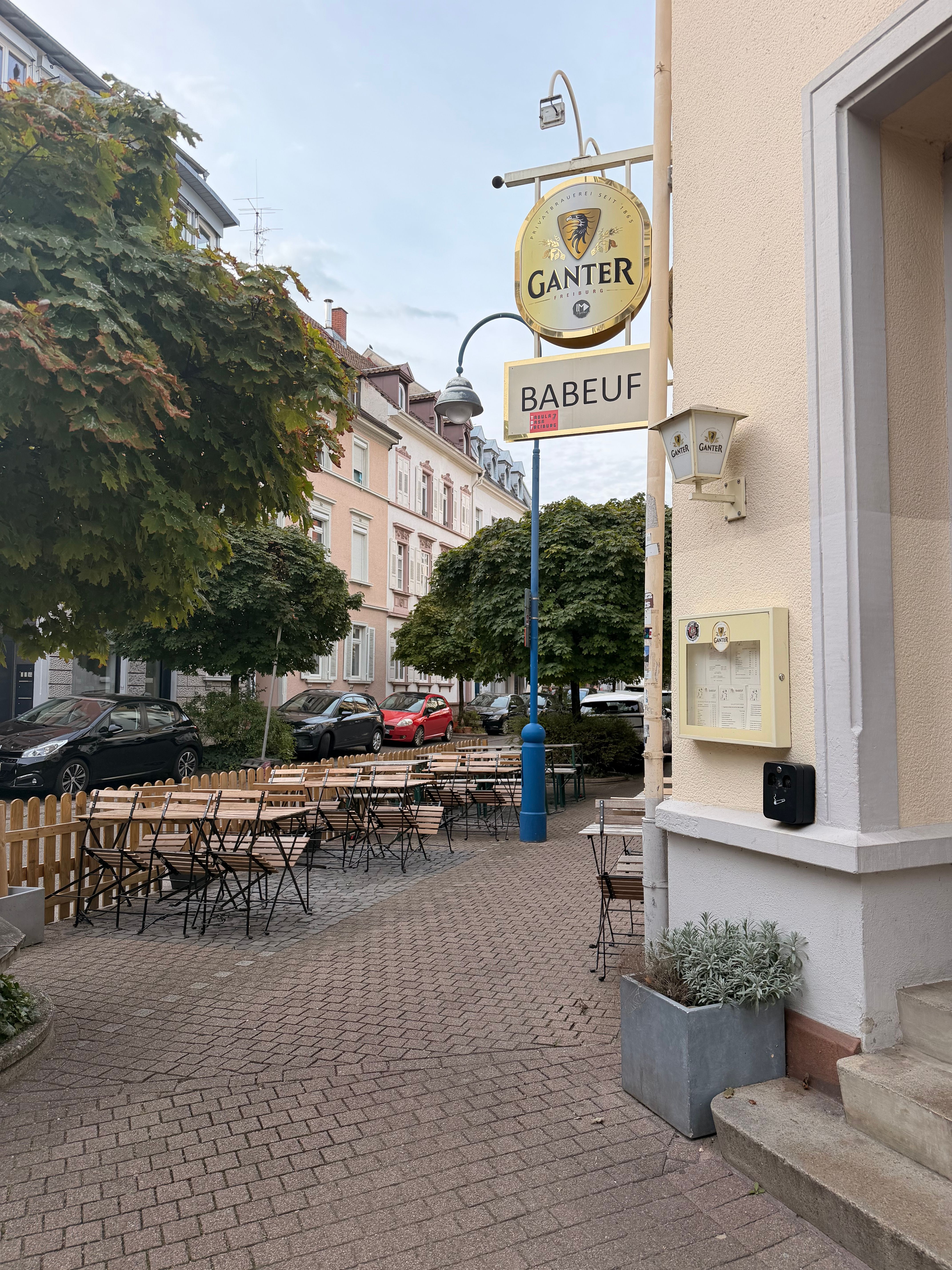 Straßenszene mit dem Ganter-Schild und dem Lokal Babeuf in Freiburg, leeren Außentischen und Wohnhäusern unter Bäumen.