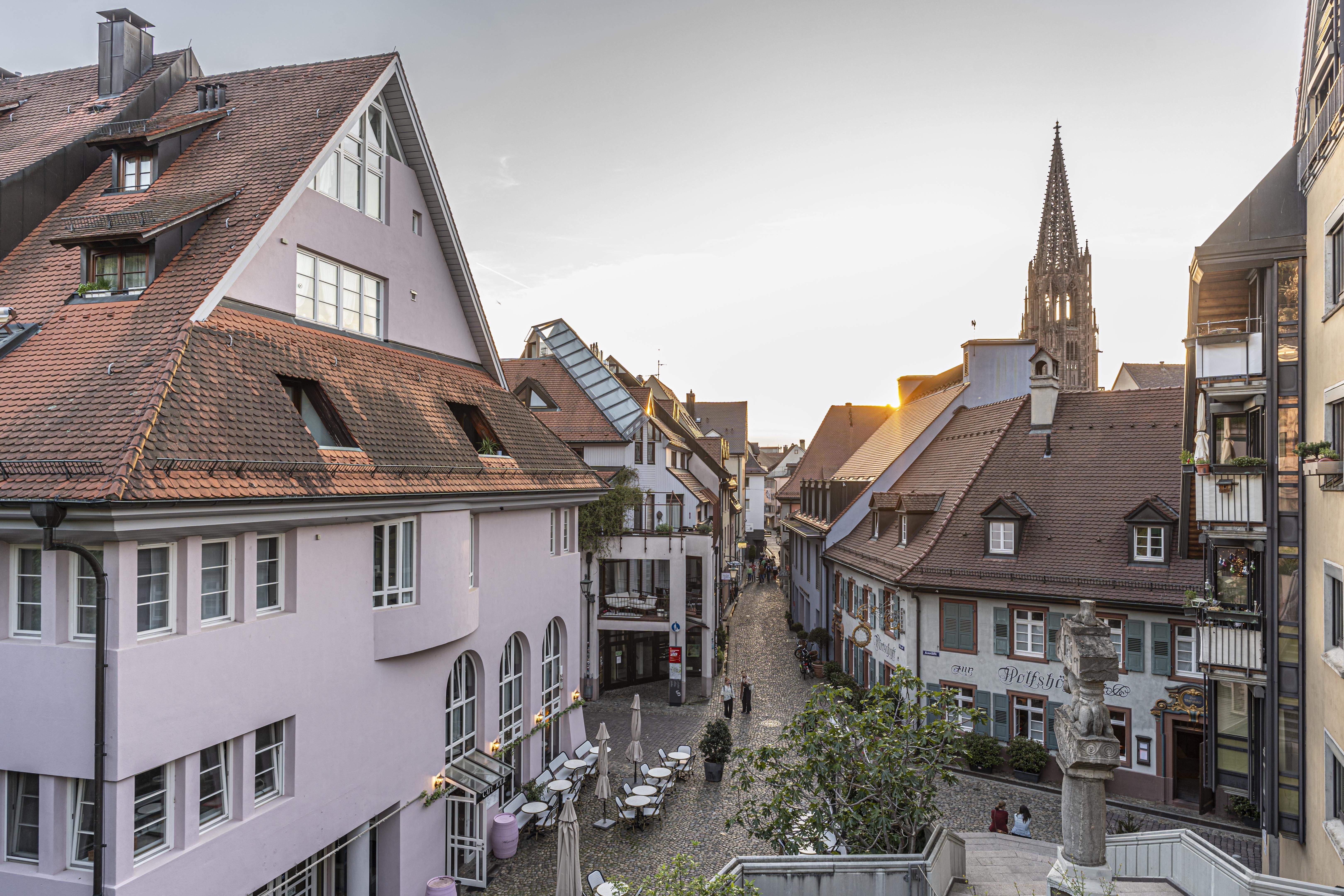 Historische Straße mit Fachwerkhäusern, Cafés und einem Kirchturm im Hintergrund bei Sonnenuntergang. Kopfsteinpflaster und gemütliche Atmosphäre.