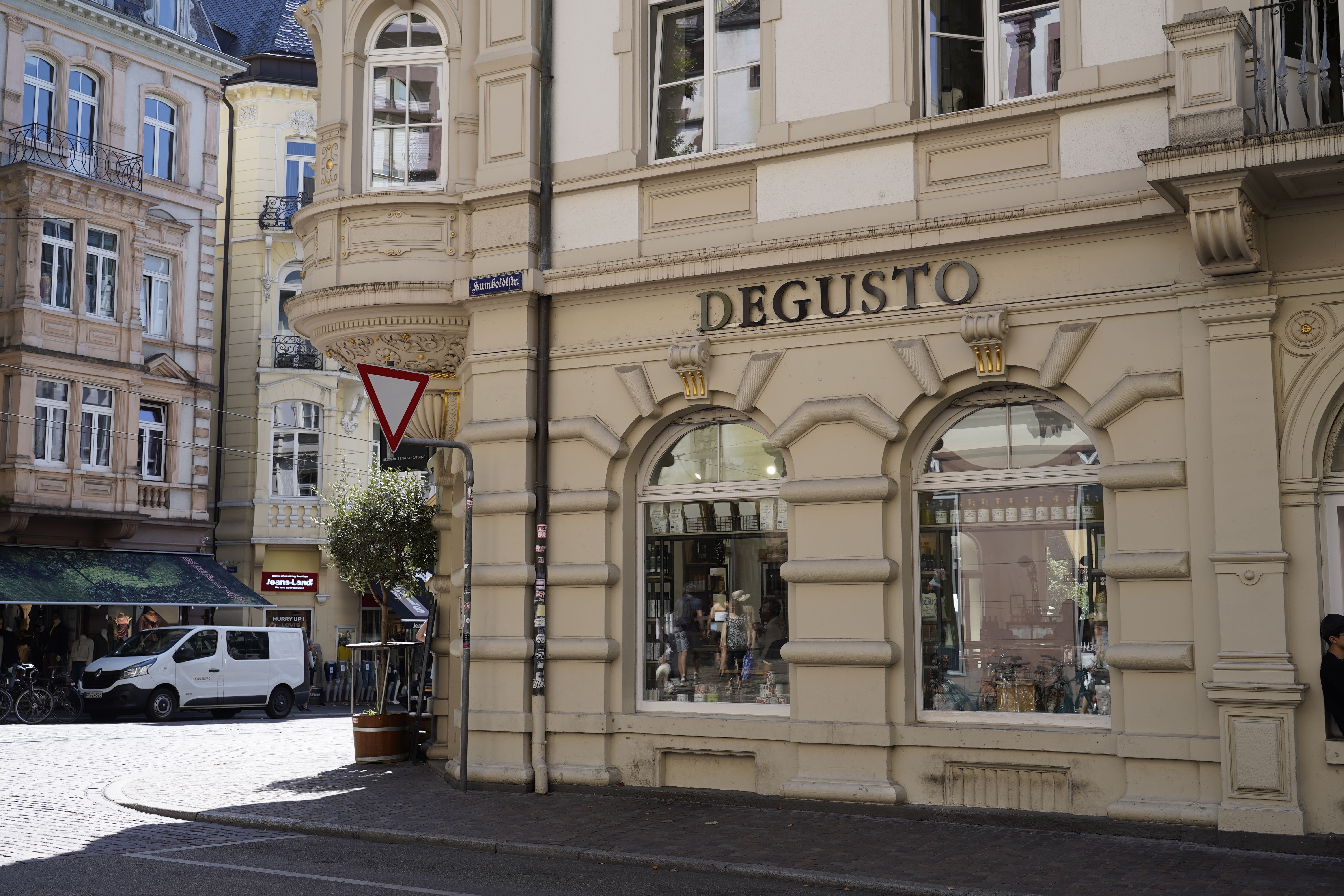 Eckgebäude mit der Aufschrift 'DEGUSTO' in einer belebten Straße, umgeben von historischen Gebäuden. Ein Lieferwagen und ein Verkehrsschild sind sichtbar.