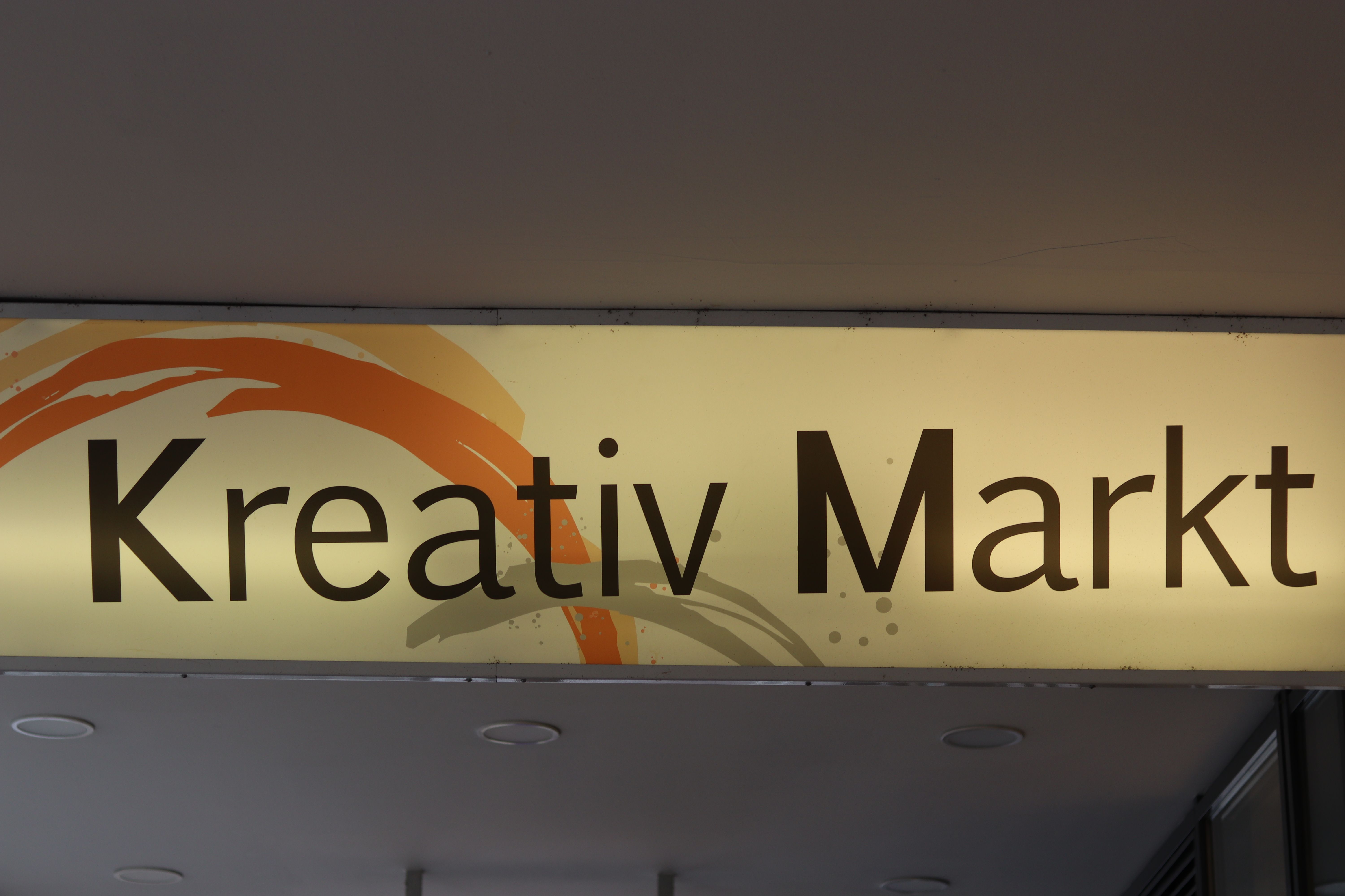 Ein beleuchtetes Schild mit der Aufschrift 'Kreativ Markt' in dunkler Schrift auf hellem Hintergrund.