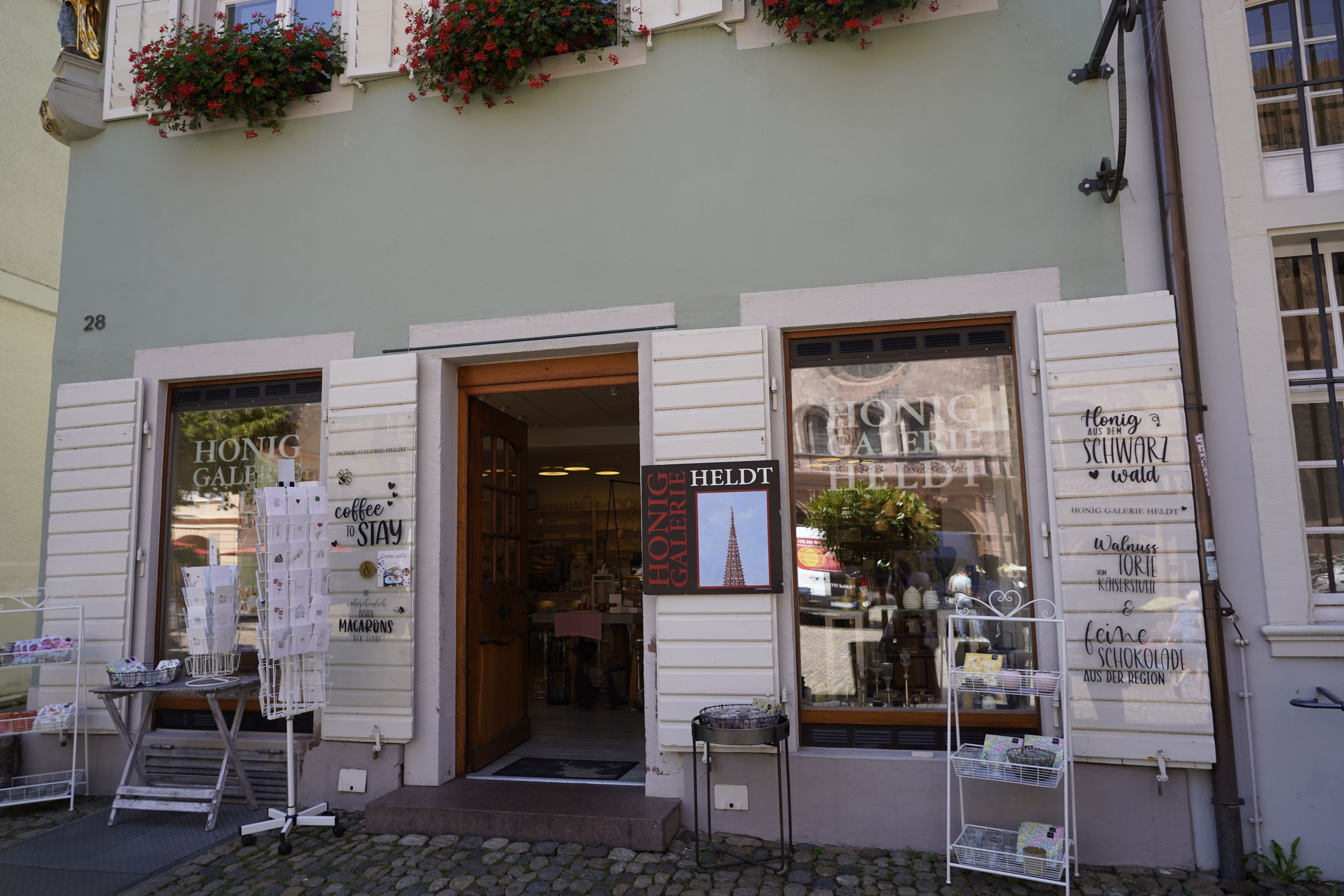 Ein Laden namens Honig Galerie Heldt mit Schaufenstern, Blumen und Dekorationen. Schilder werben für Kaffee, Macarons und regionale Produkte.