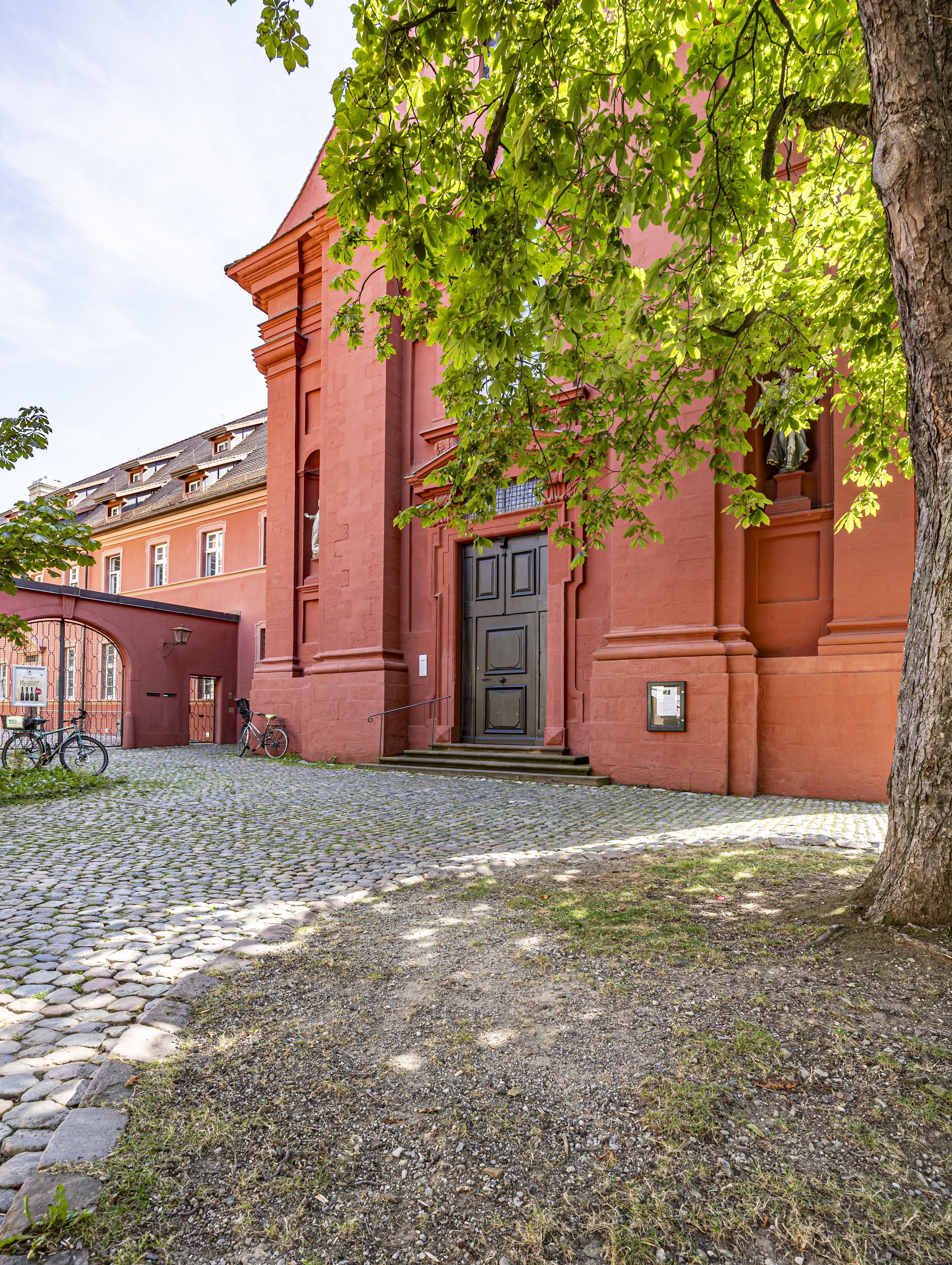 Eingang des Adelhauser Klosters an der Kirche in Freiburg, rote Fassade, Kopfsteinpflaster, Fahrräder, Baum im Vordergrund, sonniger Tag.