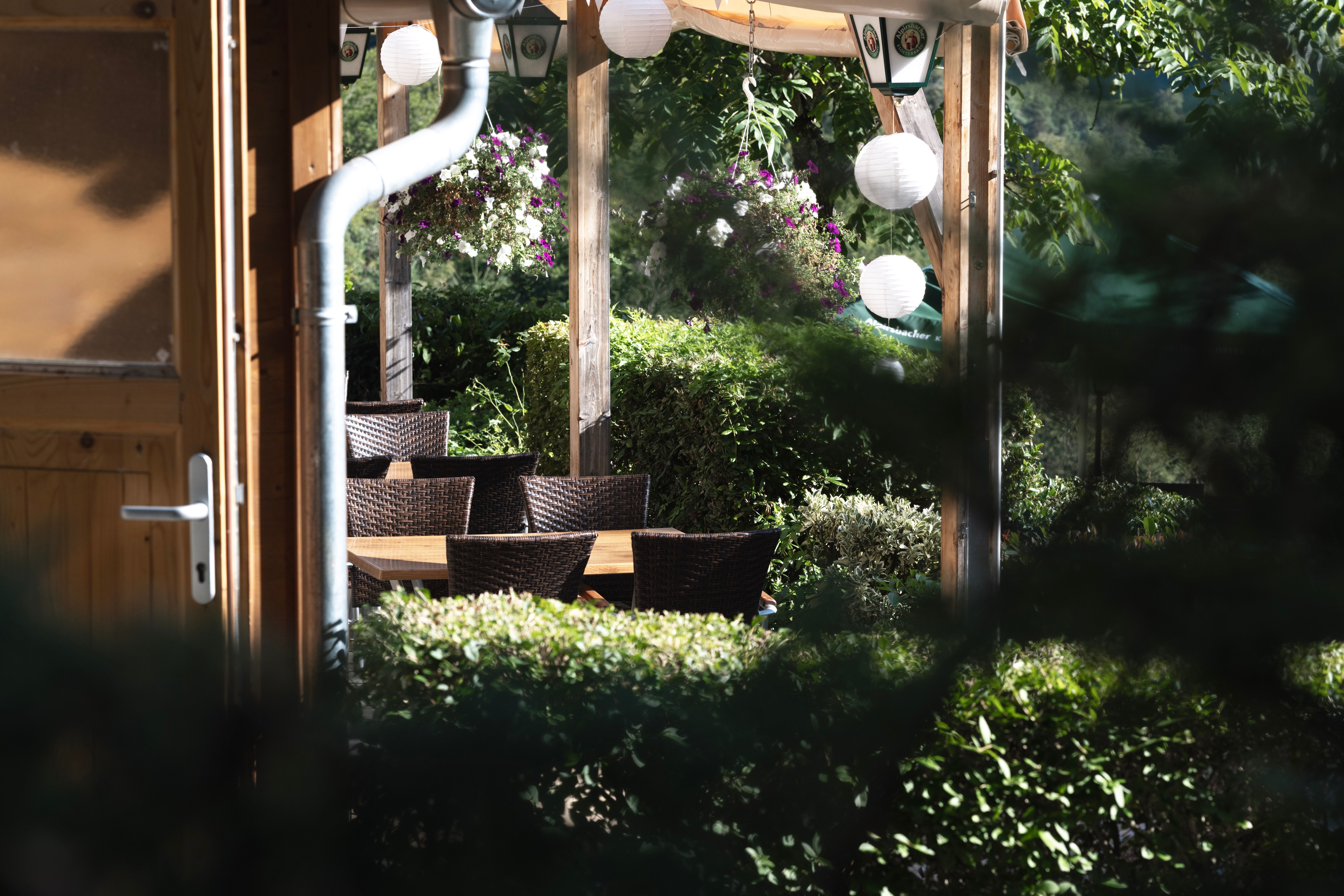 Gemütlicher Außenbereich eines Restaurants mit Holztischen, Korbstühlen und hängenden Blumen, umgeben von grüner Vegetation.