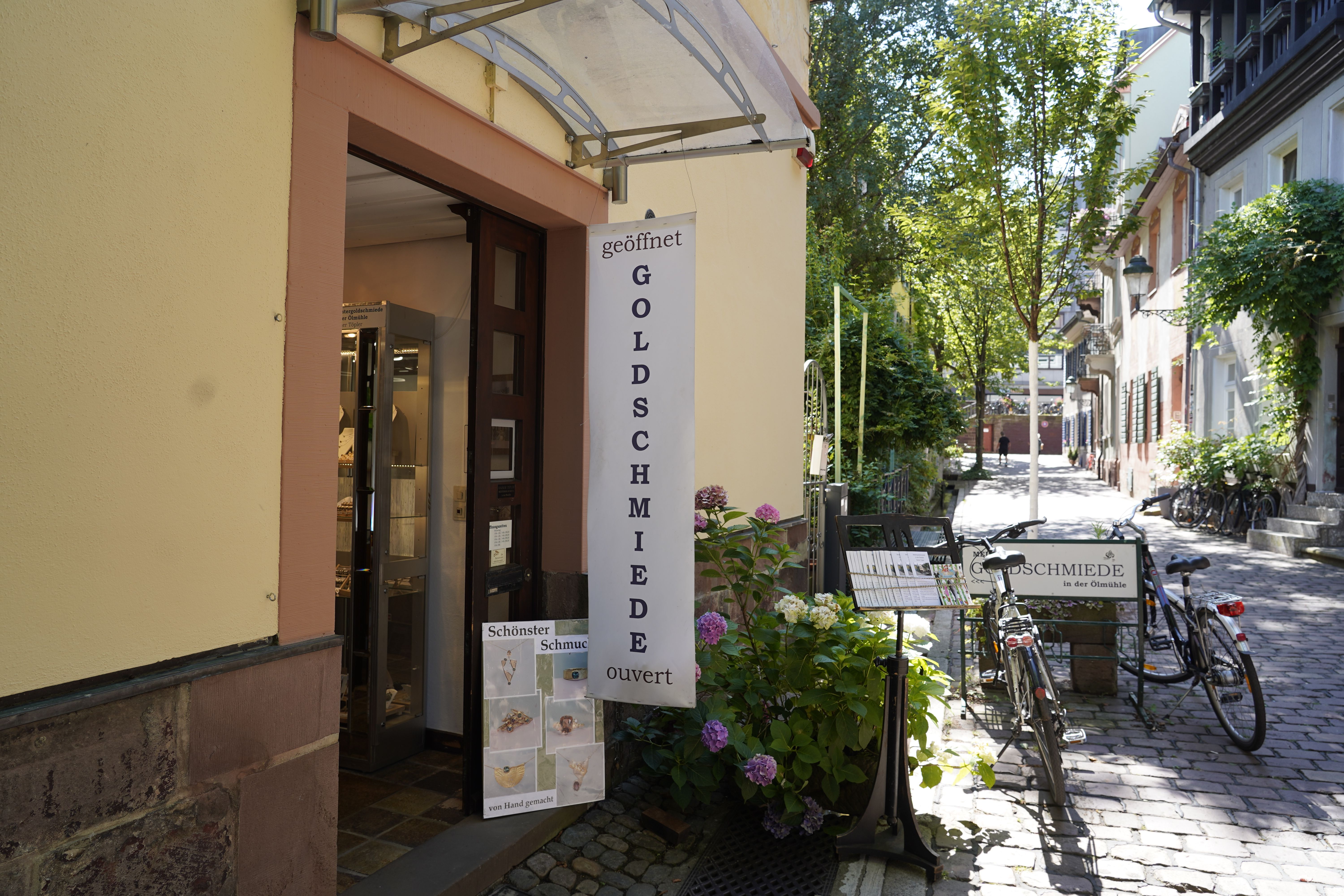 Eingang einer Goldschmiede mit Schild 'geöffnet'. Vor dem Laden stehen Fahrräder und Pflanzen. Die Straße ist gepflastert und von Bäumen gesäumt.
