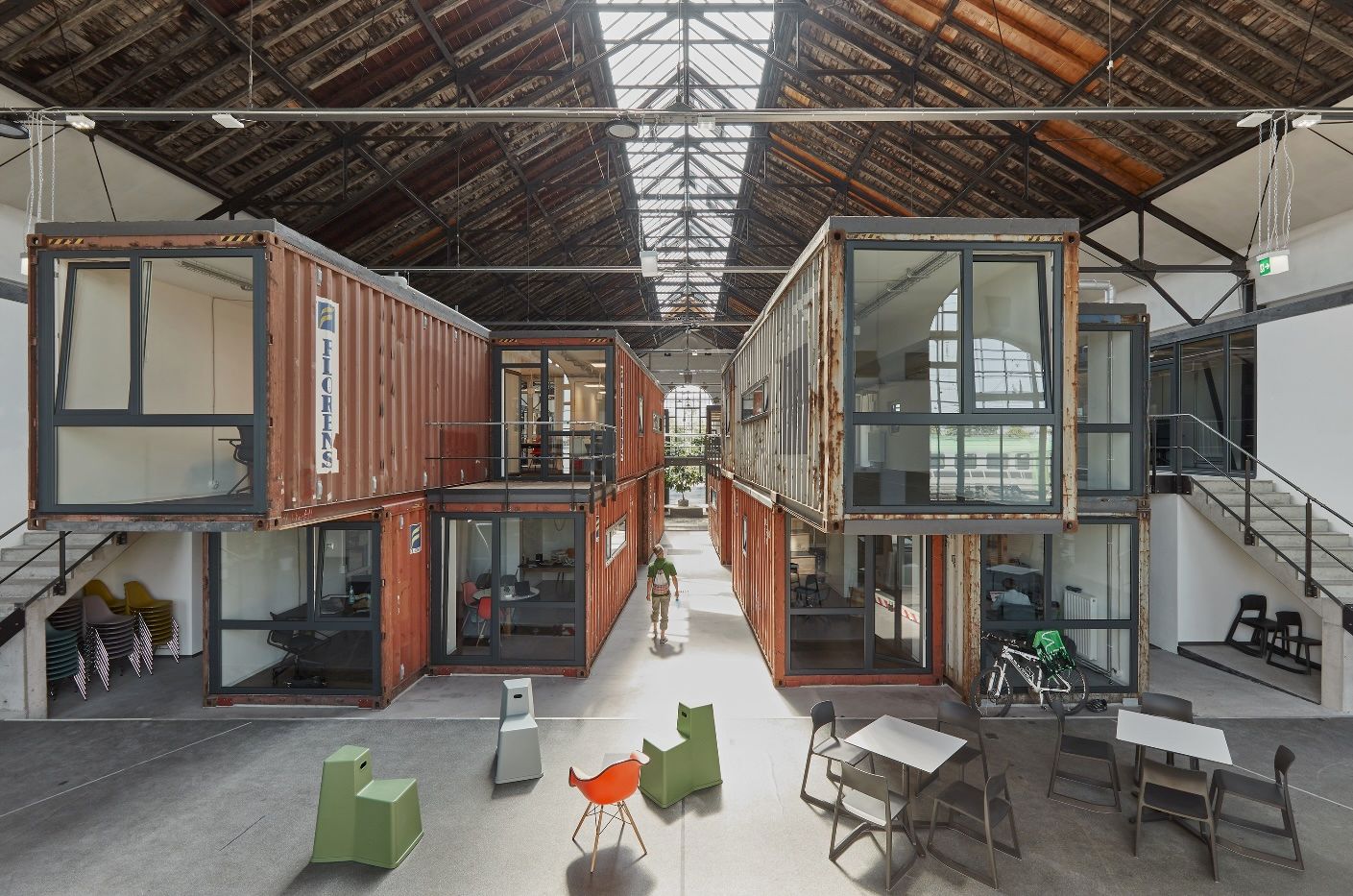 Moderne Arbeitsumgebung in einer Halle mit Containern als Büros. Helle, offene Gestaltung mit Sitzgelegenheiten und einem Fahrrad im Vordergrund.