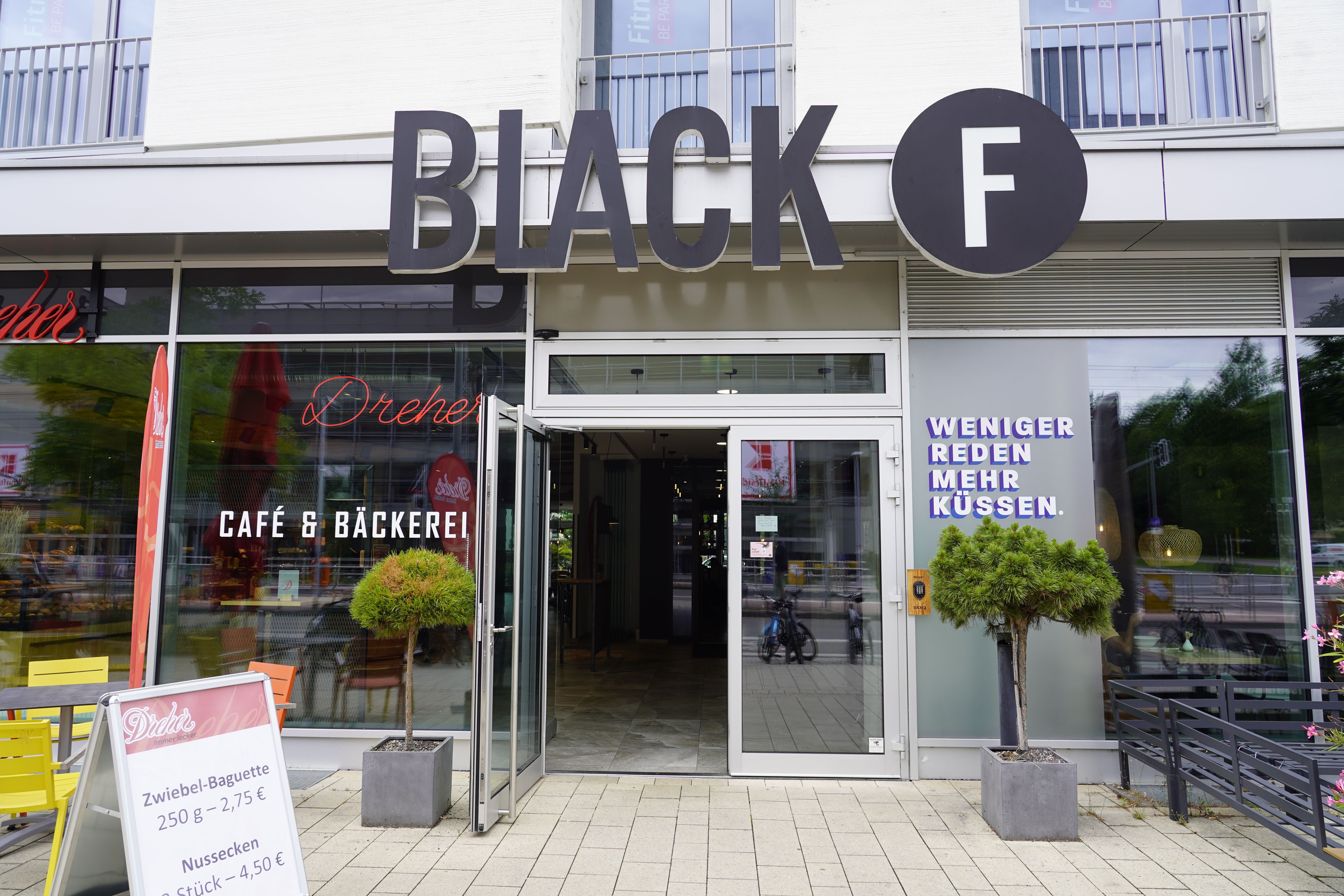 Außenansicht des Black F House mit großen Fenstern, offenen Türen und Pflanzen. 