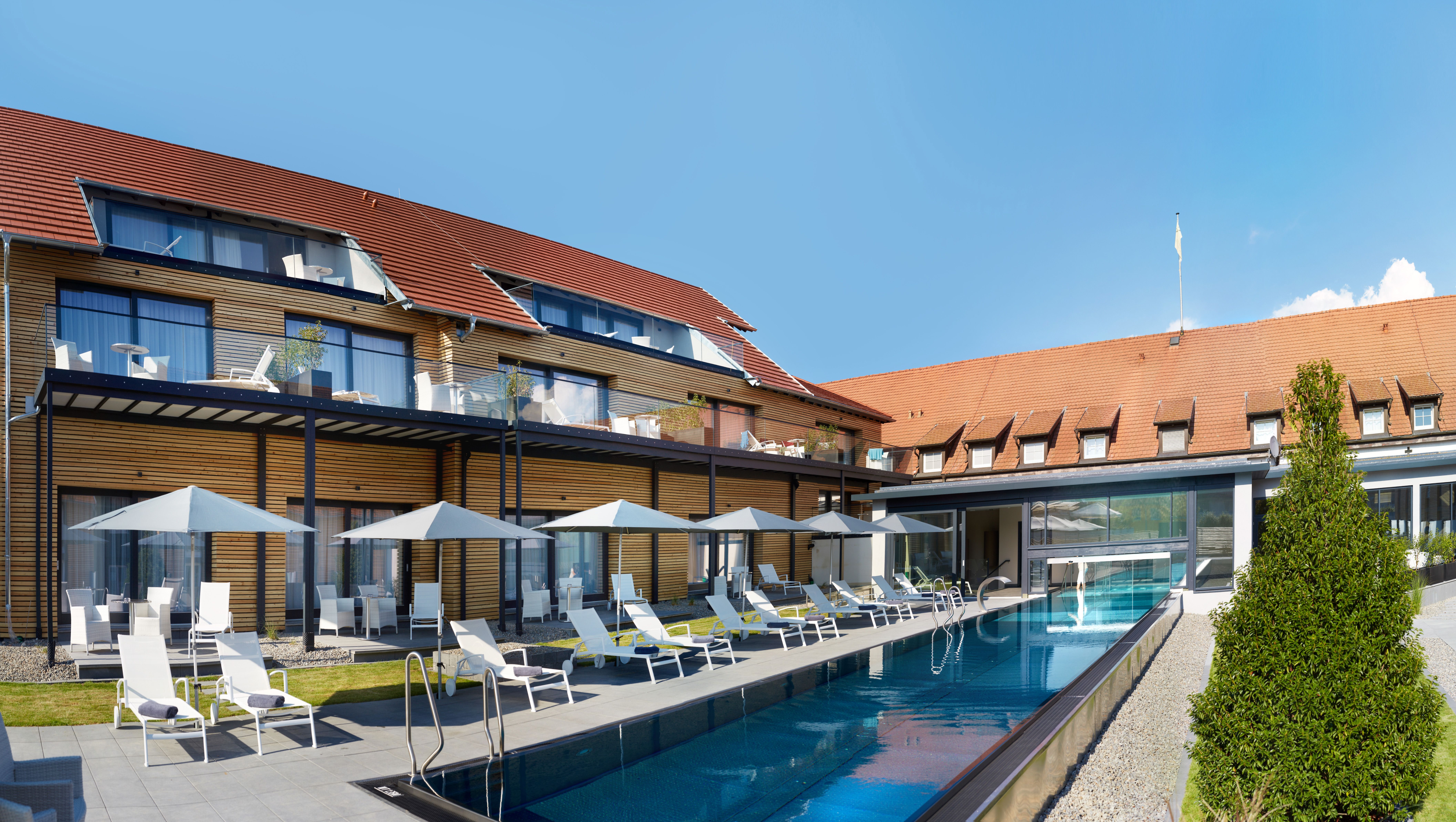 Modernes Hotel mit Pool, Liegestühlen und Sonnenschirmen. Holzfassade und rote Ziegeldächer, blauer Himmel im Hintergrund.