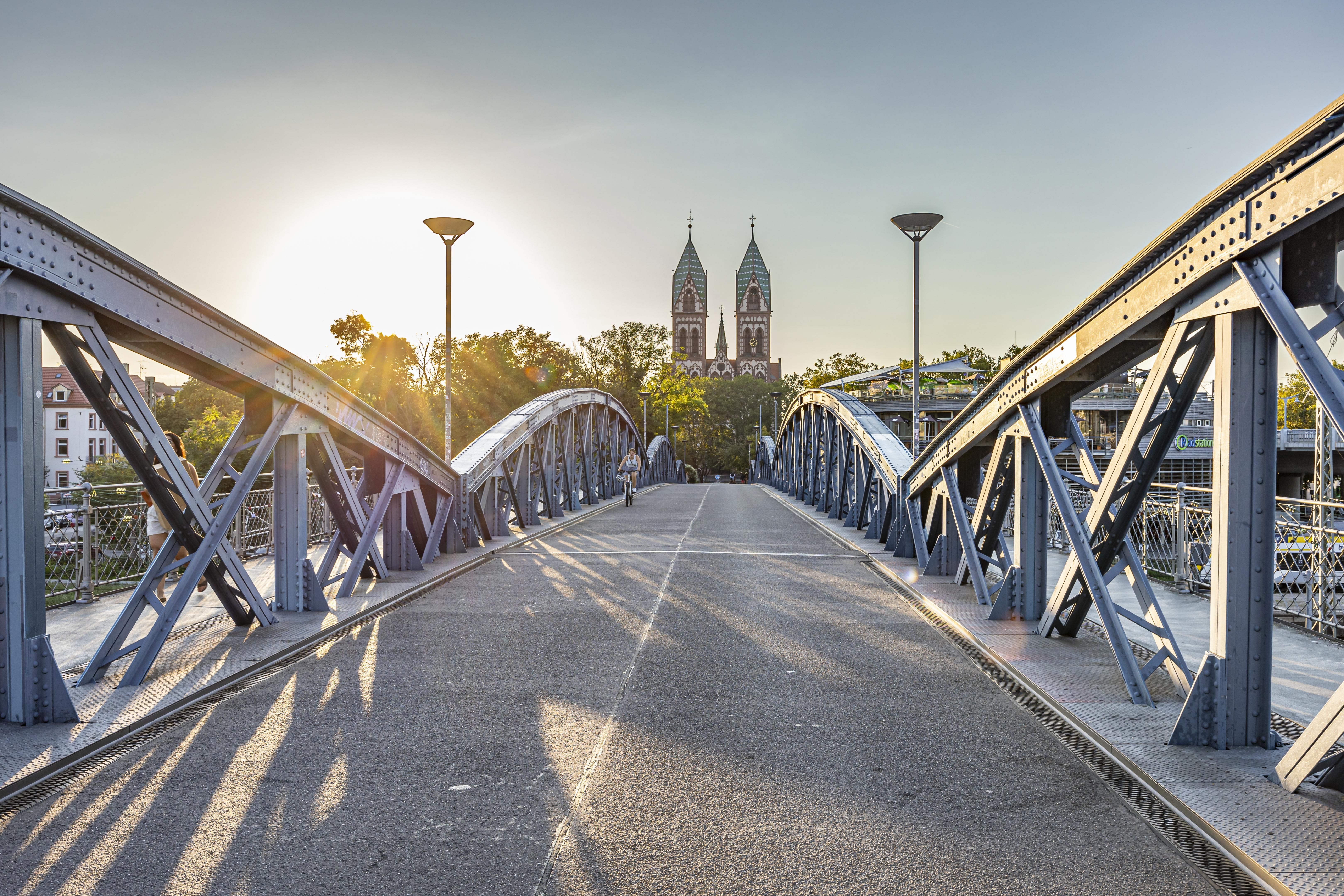 Die Wiwili-Brücke in Freiburg bei Sonnenuntergang, mit einer Kirche im Hintergrund. Ein Radfahrer überquert die Brücke.