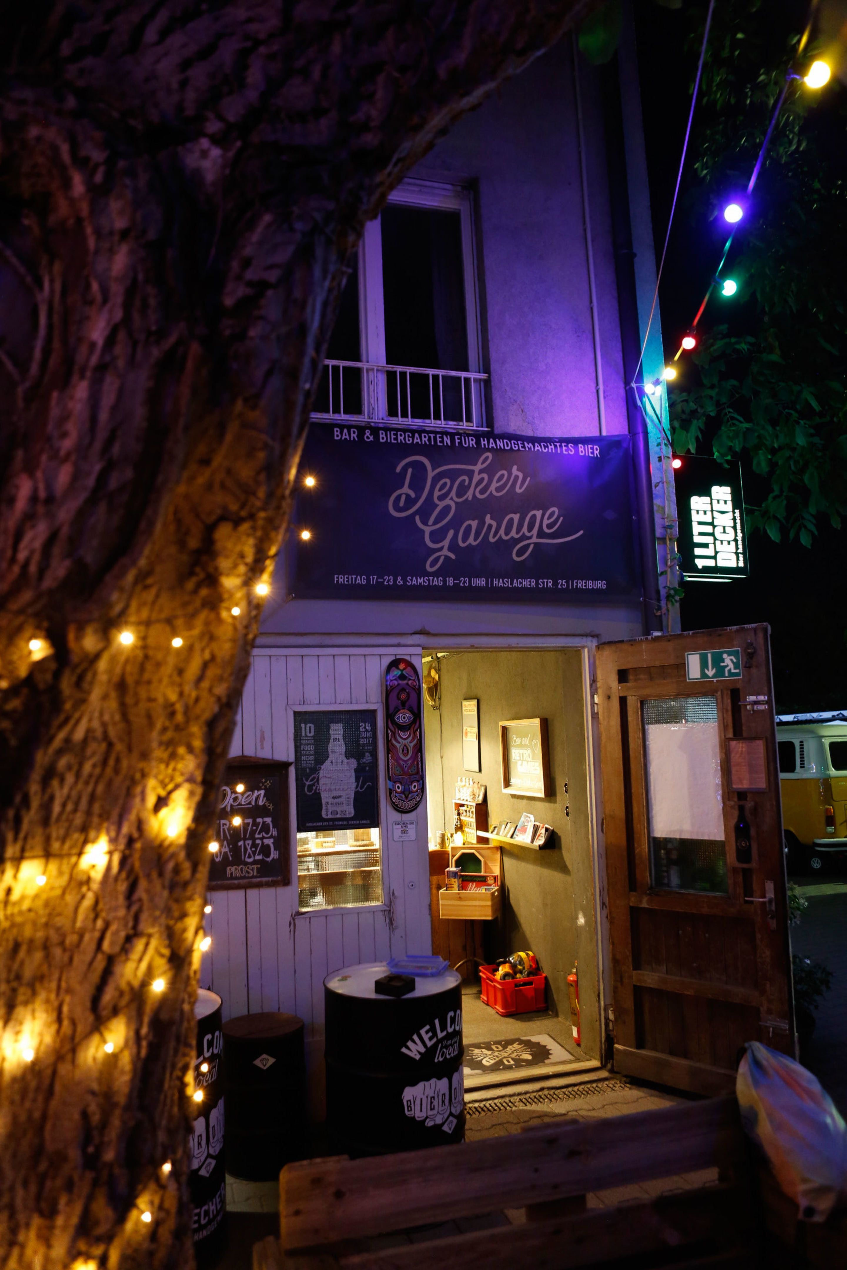 Eingang zur Decker Garage Bar in Freiburg, beleuchtet mit bunten Lichtern und Lichterketten an einem Baum bei Nacht.