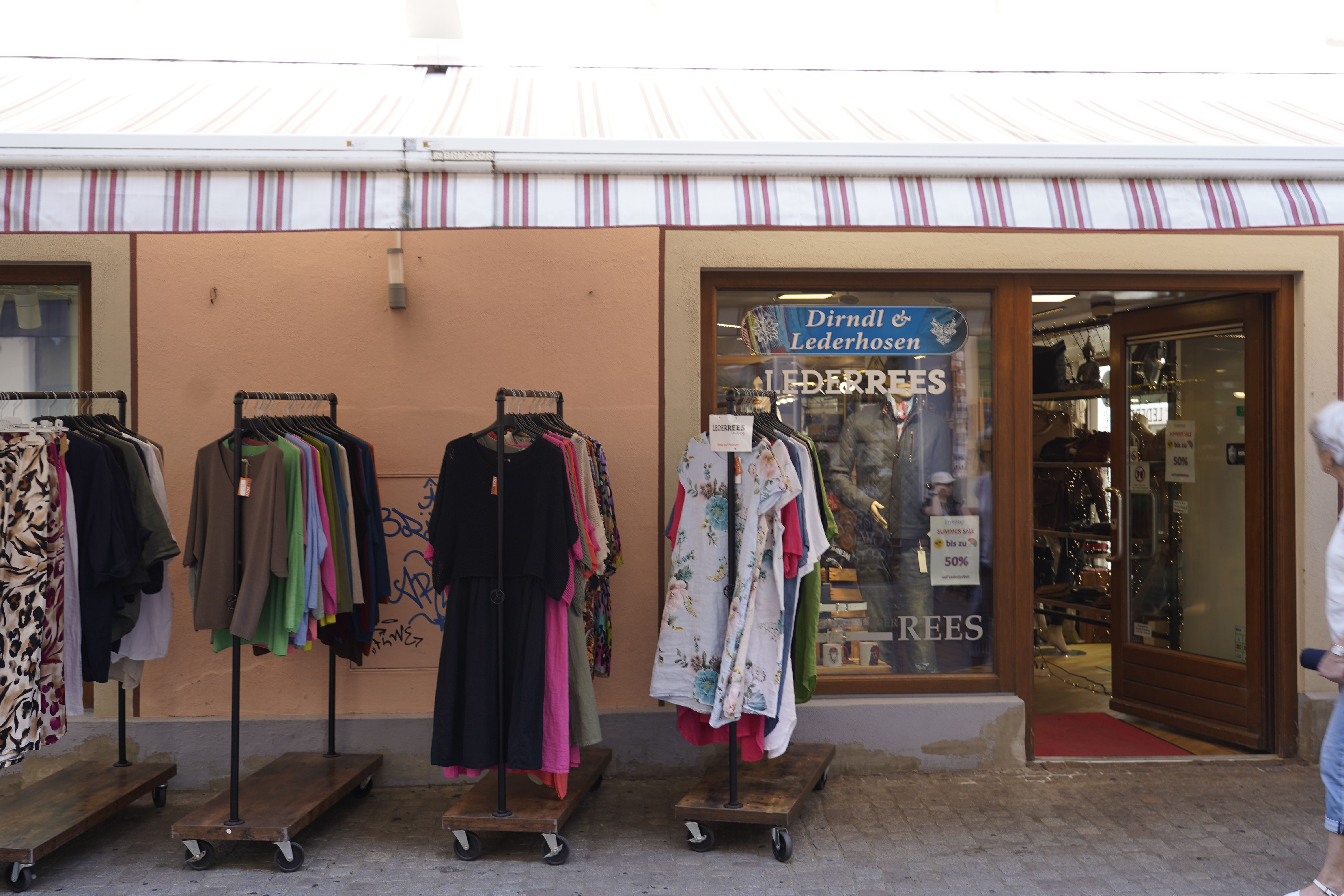 Ein Bekleidungsgeschäft in Freiburg mit Kleidungsständern draußen. Im Schaufenster steht "Dirndl & Lederhosen". Eine Person geht vorbei.