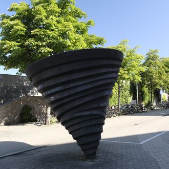 Schwarze, spiralförmige Skulptur auf einem gepflasterten Platz, umgeben von Bäumen und Fahrrädern bei klarem Himmel.
