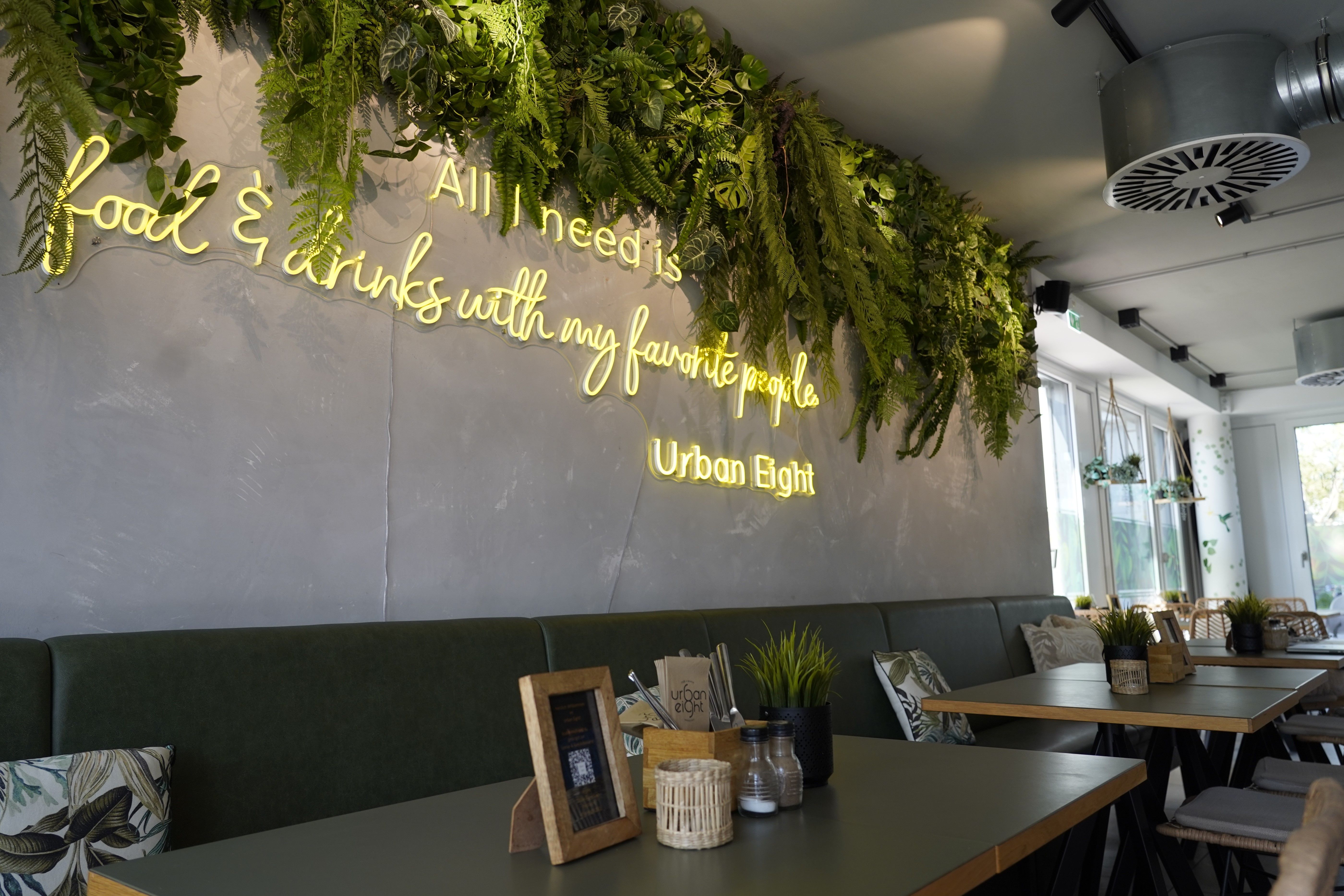 Modernes Café mit grüner Dekoration, Neon-Schriftzug "All I need is food & drinks with my favorite people." an der Wand.