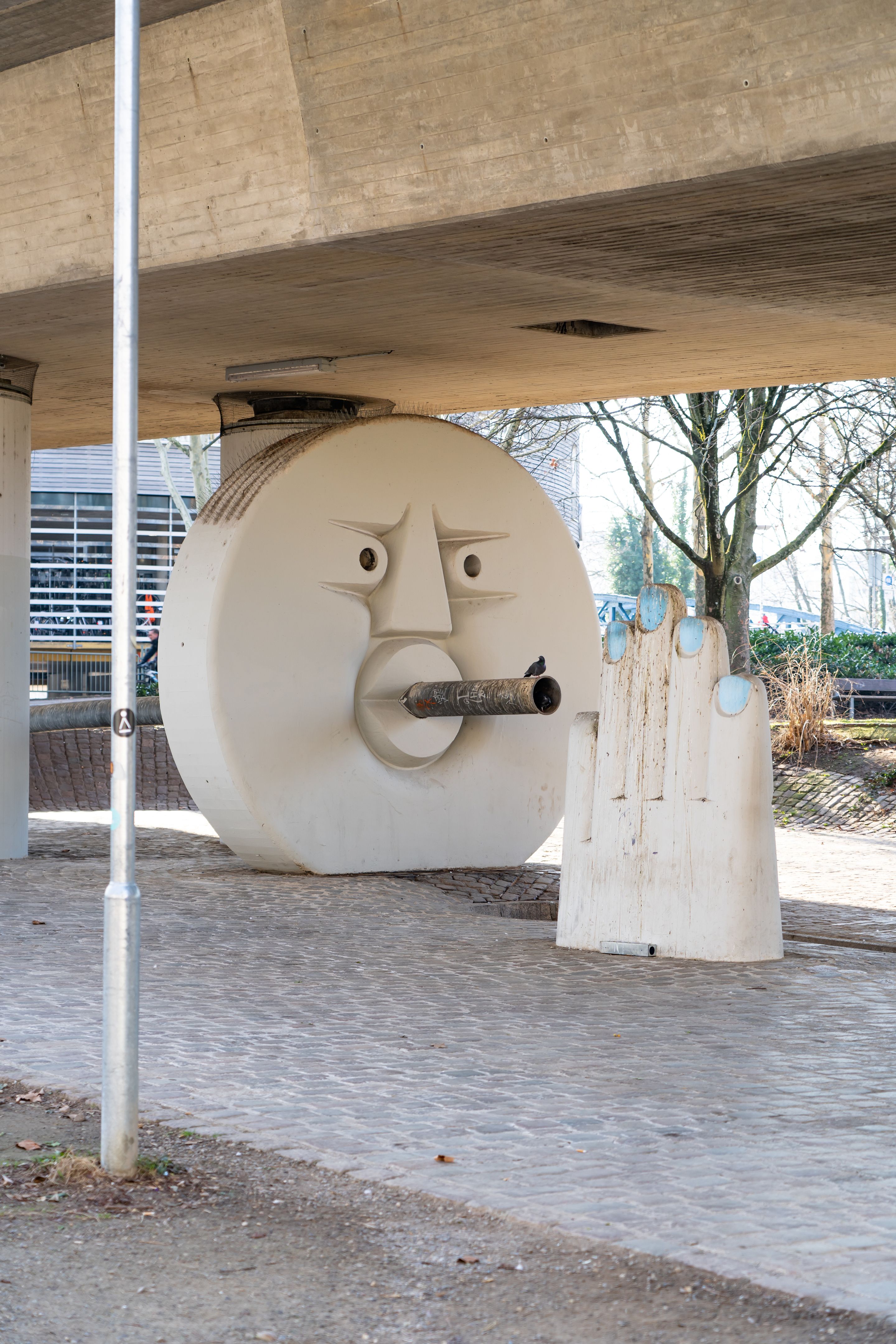 Eine weiße Skulptur mit Gesicht und Hand unter einer Brücke in Freiburg. Die Skulptur hat ein Rohr als Mund.