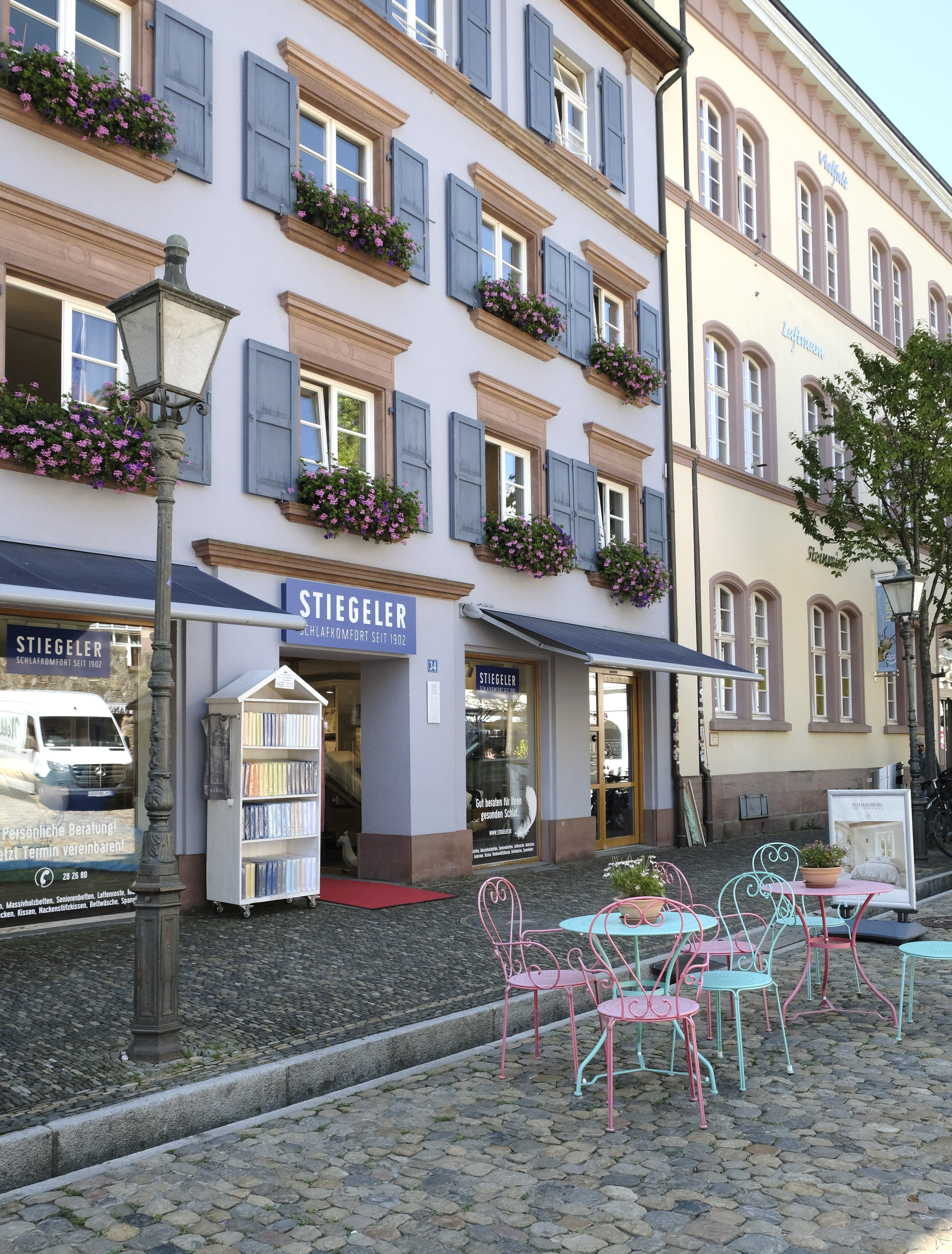 Gebäude mit 'Stiegeler Schlafkomfort', blauen Fensterläden, Blumen. Bunte Tische und Stühle auf Kopfsteinpflaster davor.