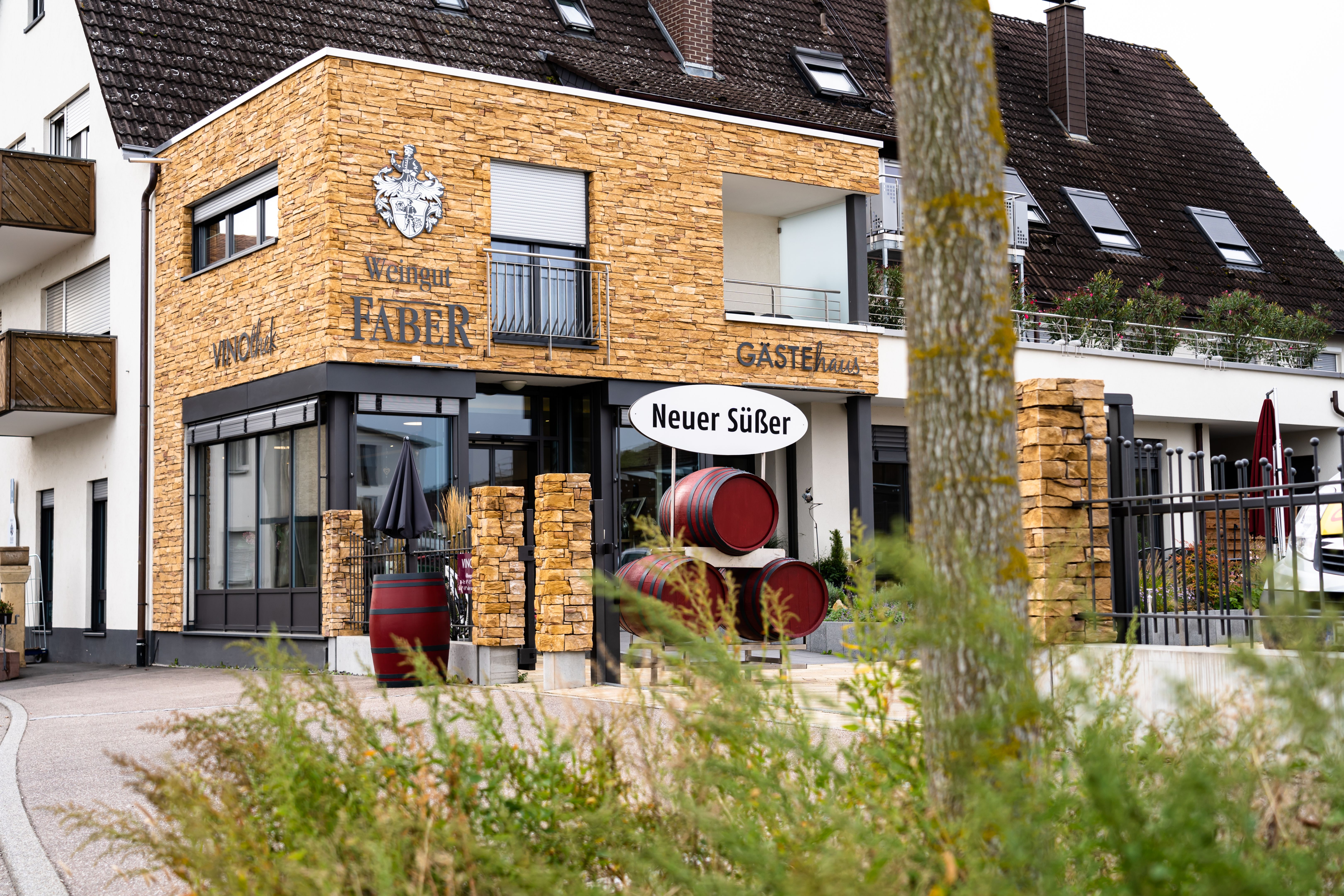 Weingut Faber mit Steinverkleidung, Weinfässern und Schild 'Neuer Süßer'. Gebäude mit moderner Architektur und Gästehaus.